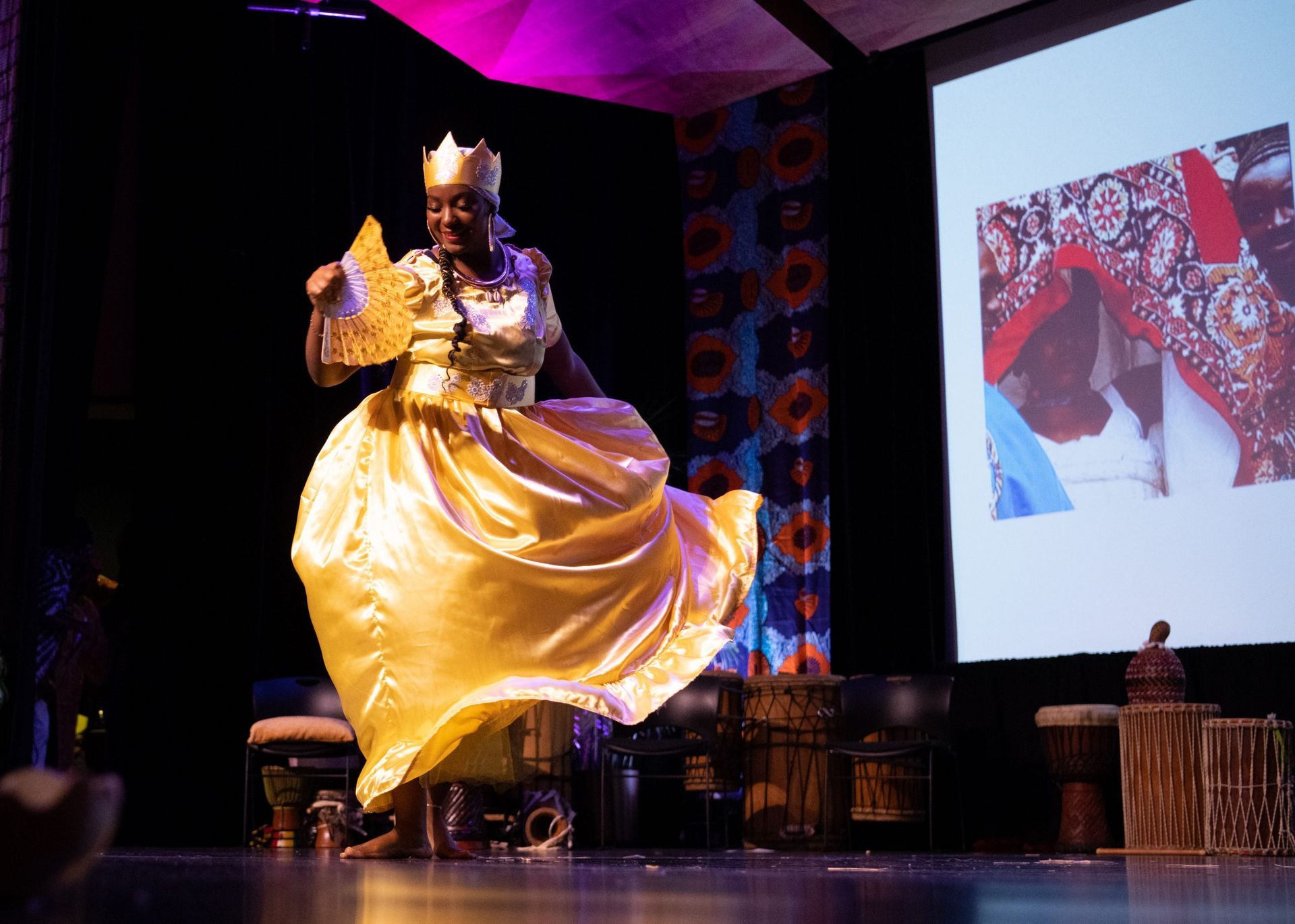 Woman in gold dress dances on stage, holding fan, wearing crown. Background features projection screen and drums.