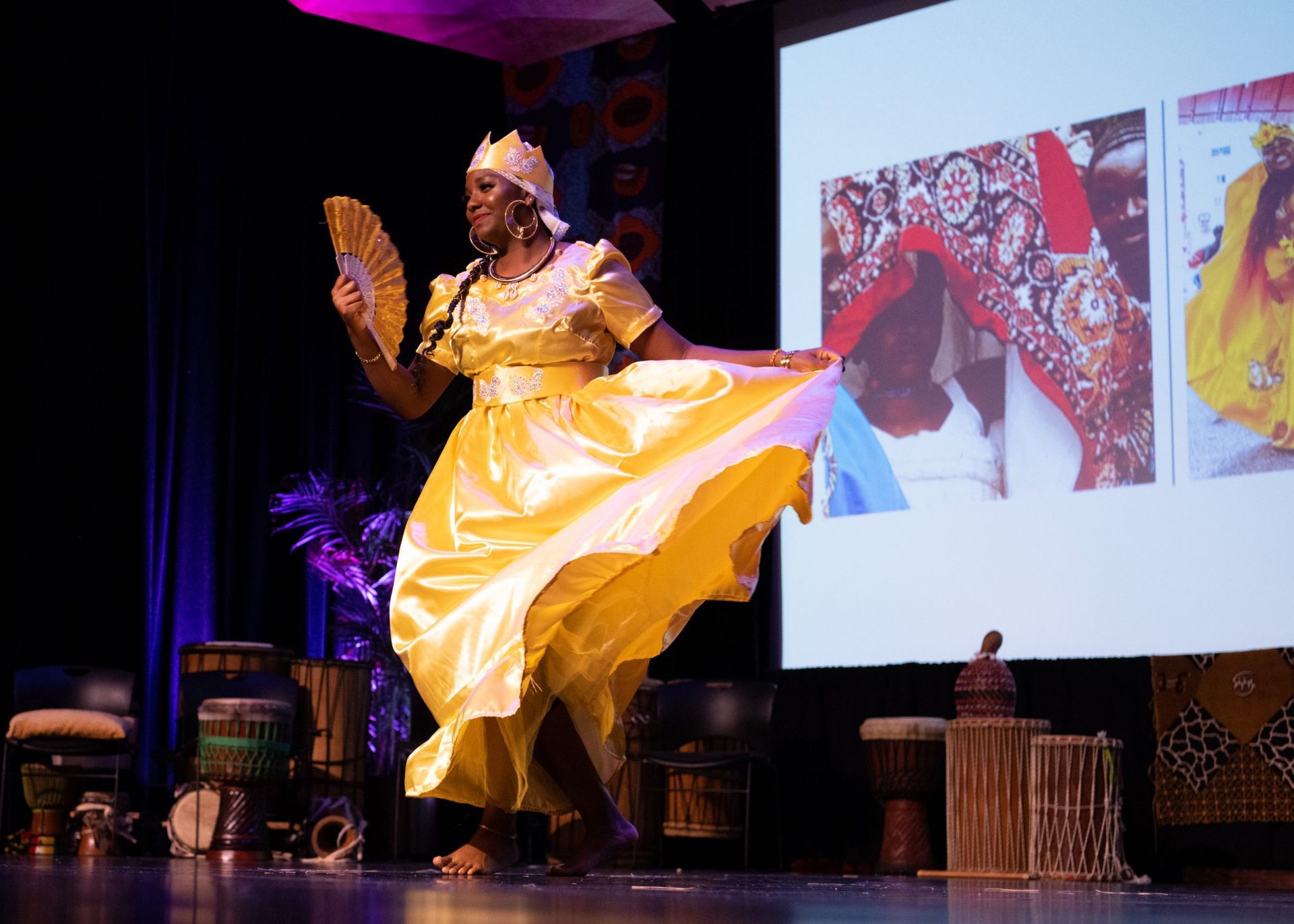 Woman in yellow dress dancing on stage, holding a fan. Behind her are drums and a screen with photos.