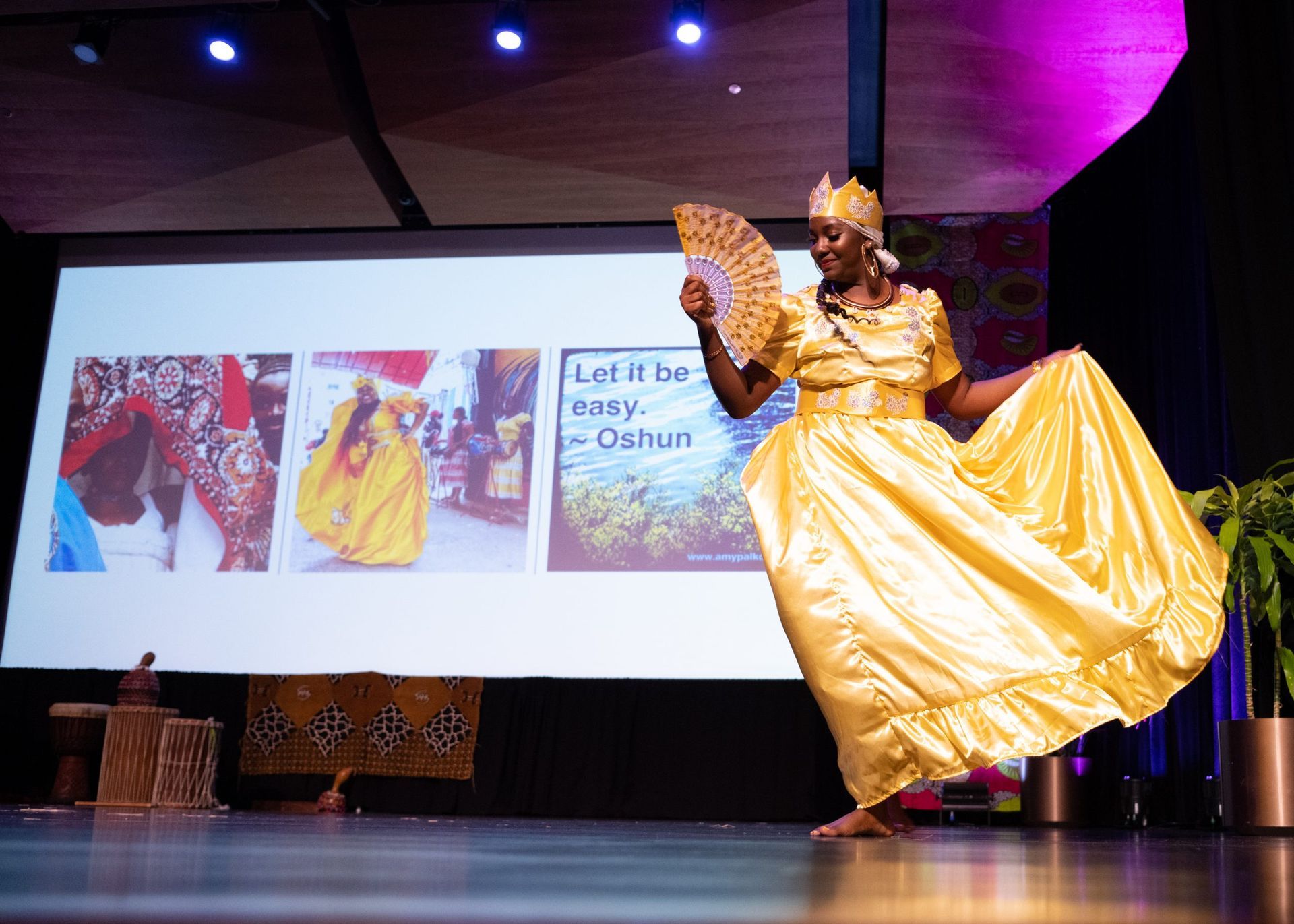Woman in gold dress dances on stage, holding a fan; backdrop includes images of Oshun.