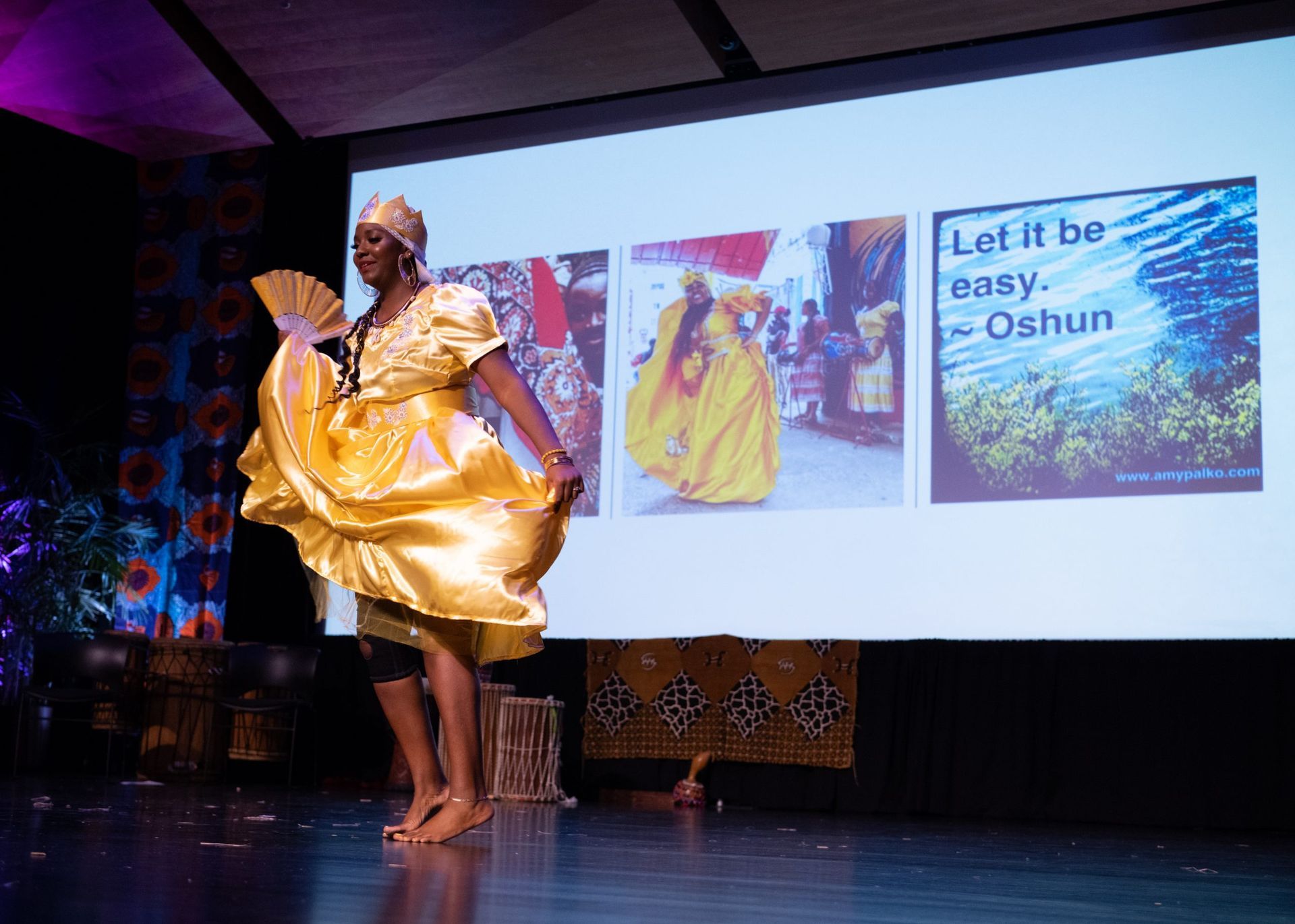 A person in a gold dress dances on stage. Behind them, a screen shows pictures and text about Oshun.