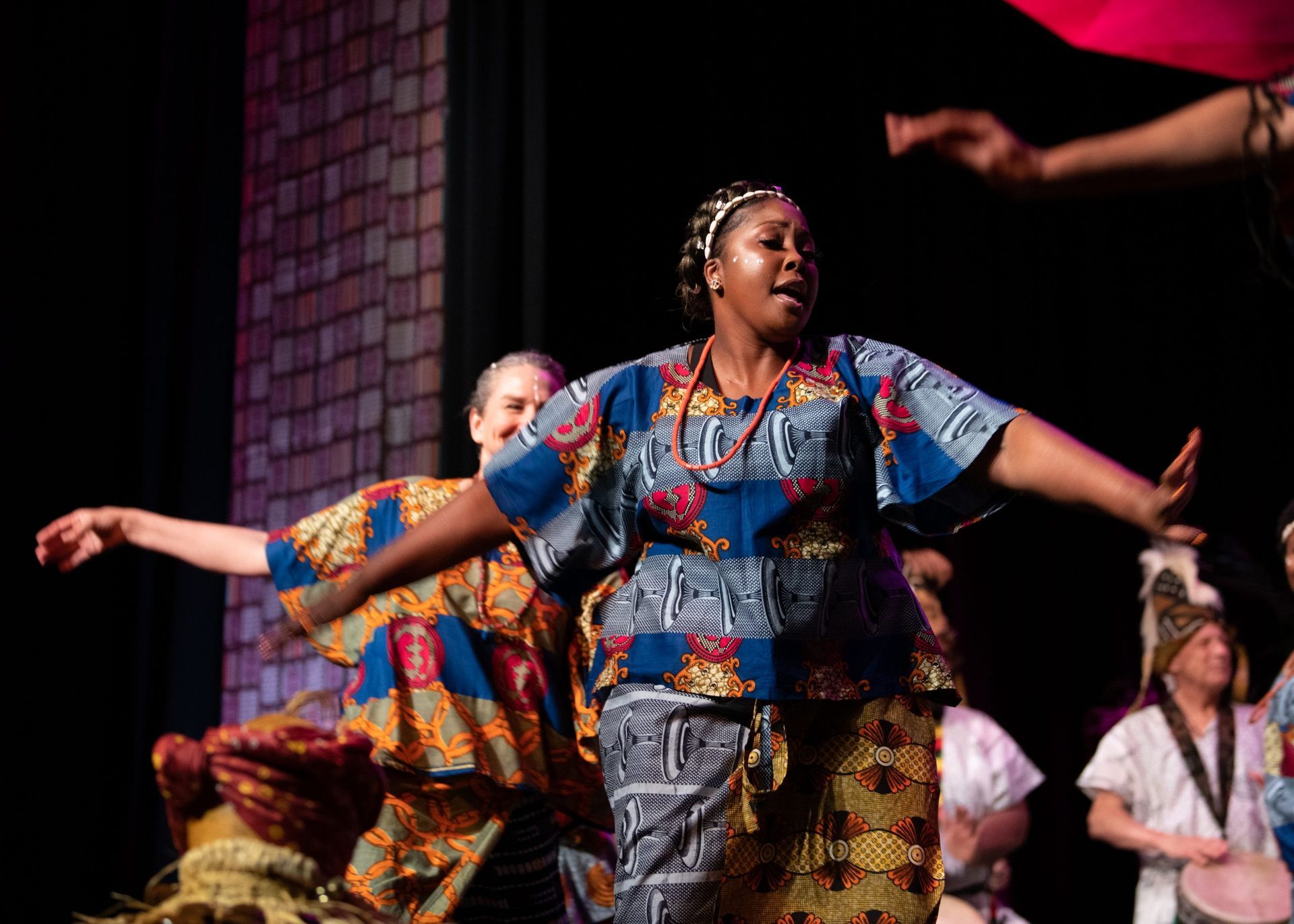 Woman in African print outfit sings on stage, arms outstretched, others dance behind her.