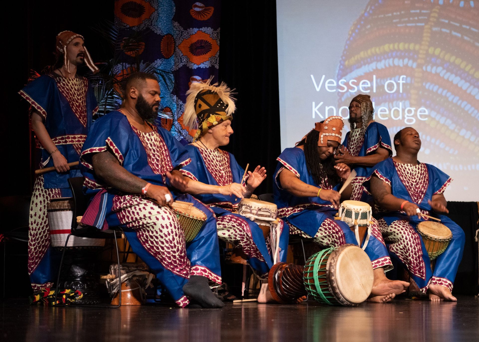 Group of people in blue and patterned outfits playing drums on stage. 