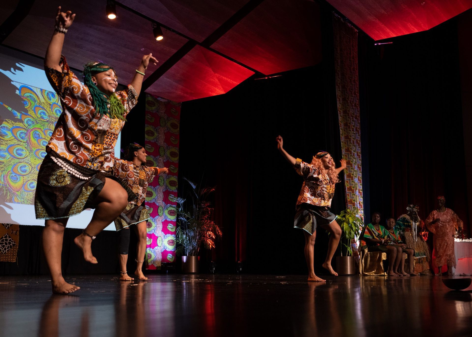 Dancers in colorful clothing perform on stage with arms raised. Red lights illuminate the background.