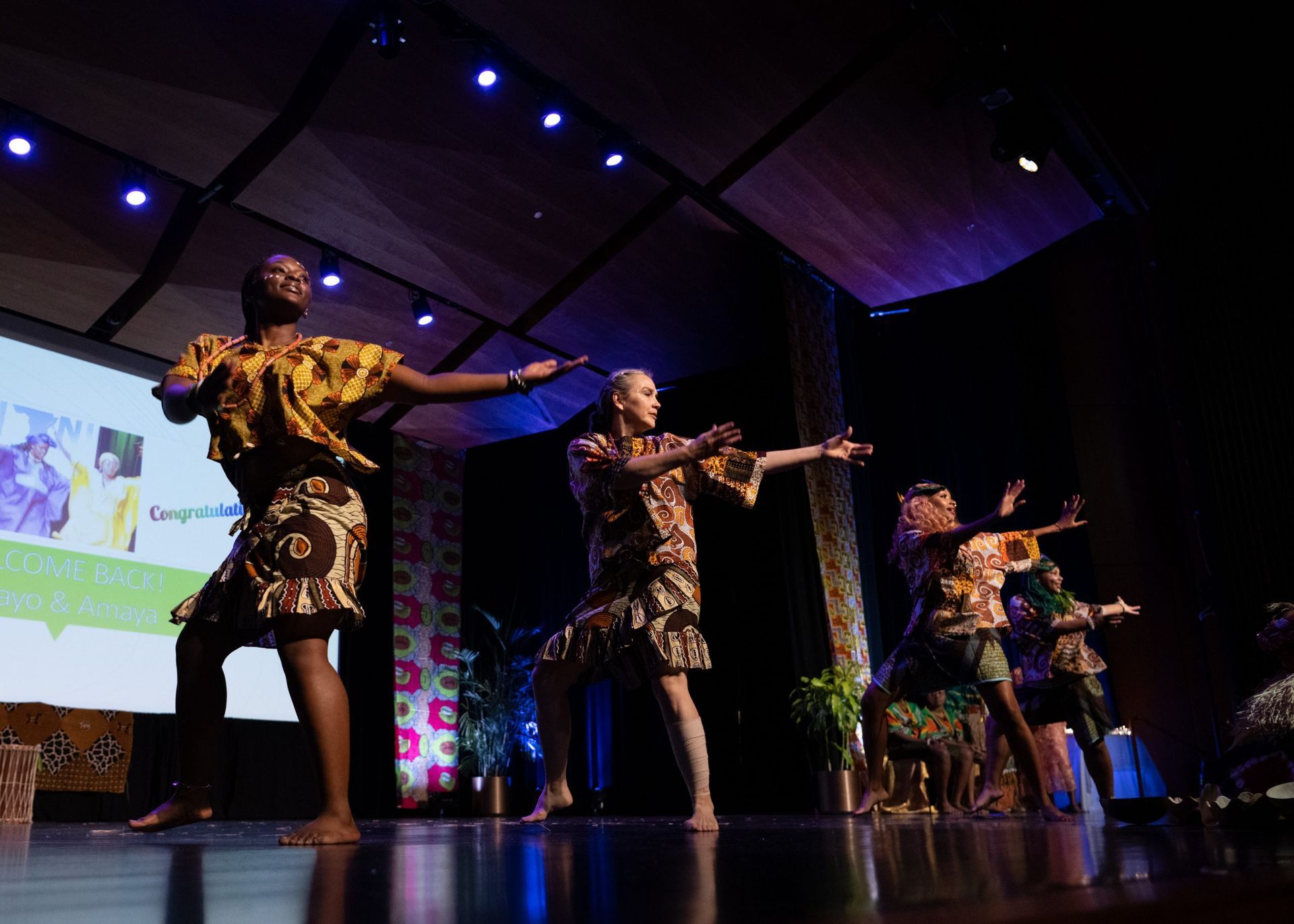 Dancers on stage wearing colorful patterned outfits perform under stage lights.