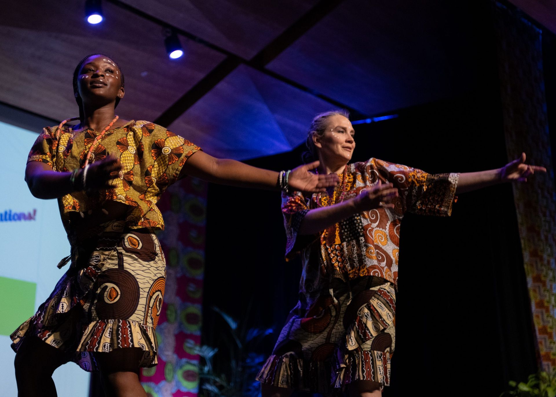 Two people in patterned outfits dance on a stage with blue and colorful lighting.