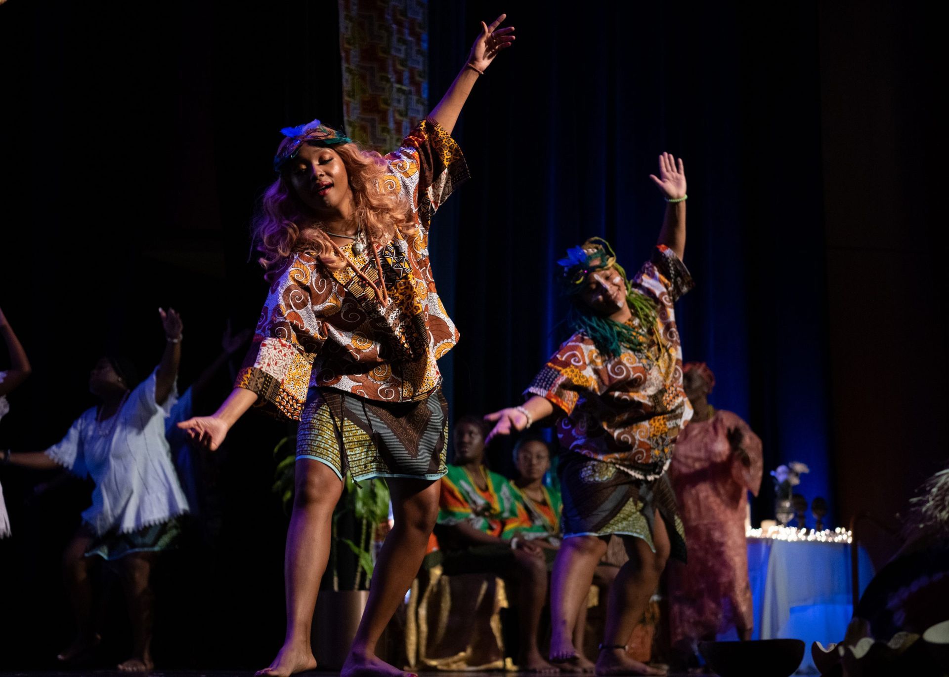 Two dancers in colorful African attire perform on stage. Others in the background.