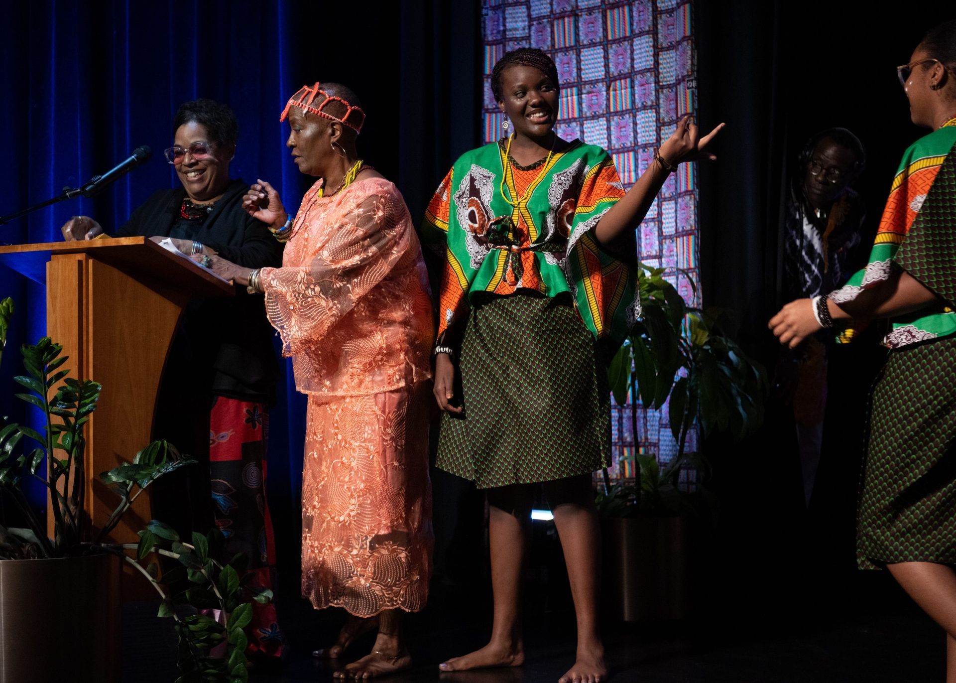 People on stage in colorful African attire; one at podium, others dancing with smiles.
