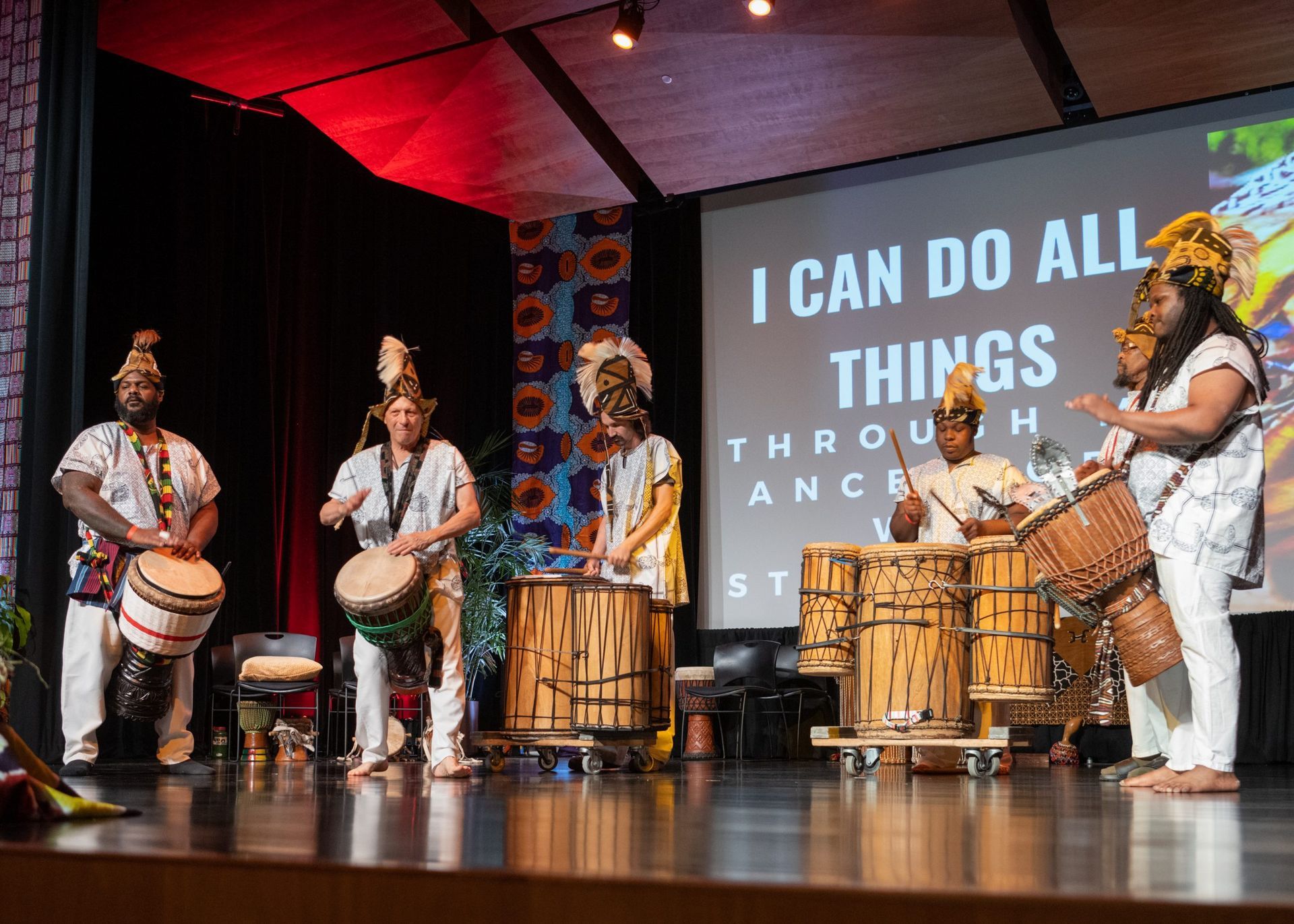 Group of musicians playing drums on a stage with a backdrop screen, all wearing white outfits with headpieces.
