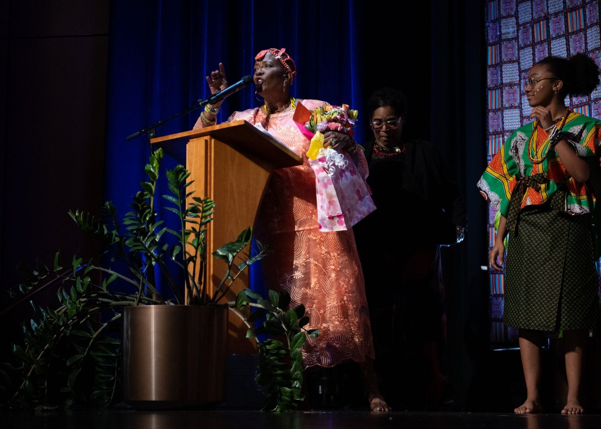 Woman speaking at a podium, receiving flowers, with two others on stage; blue background.