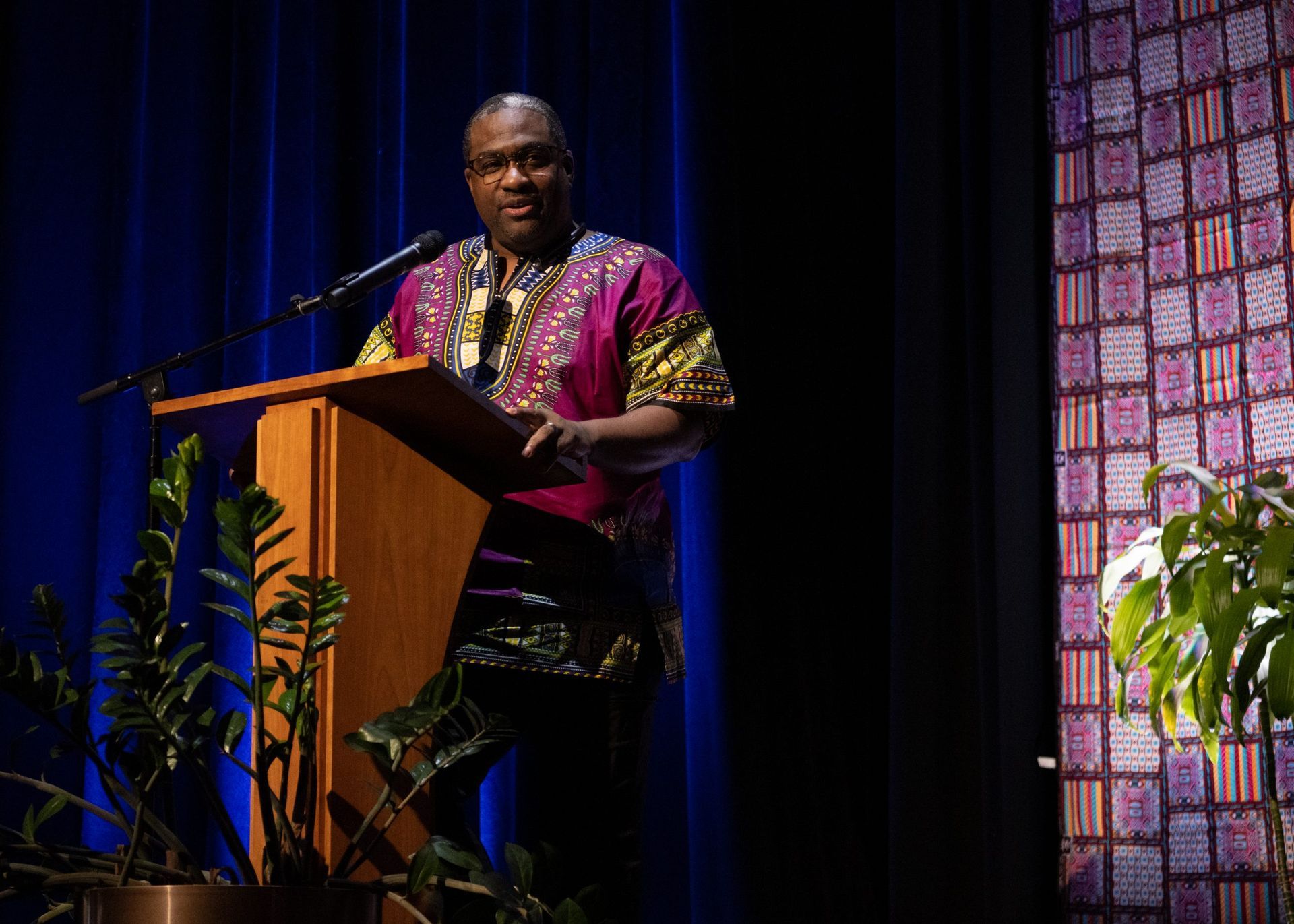 Man in colorful shirt speaks at podium with plants and patterned wall.