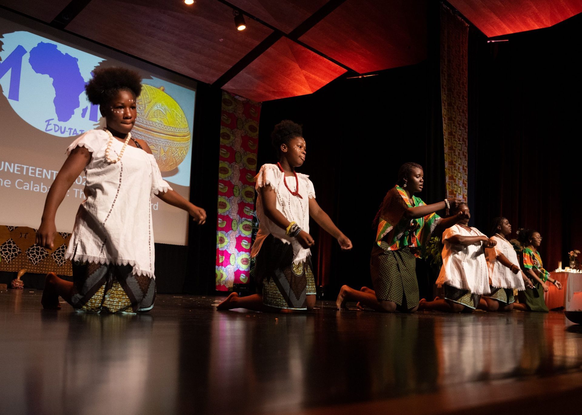 Performers in white and patterned clothes on stage, kneeling and dancing. Backstage decor, warm lighting.