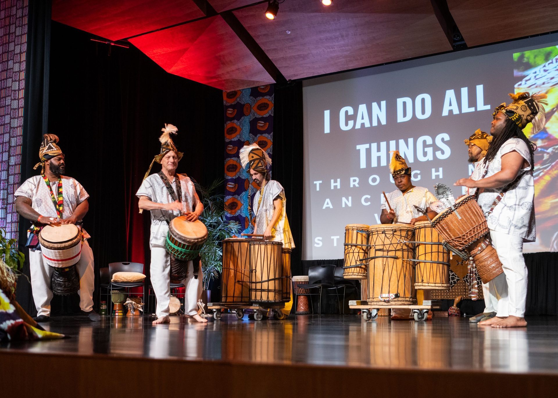 Drummers on stage performing, wearing white and patterned clothing, with text 