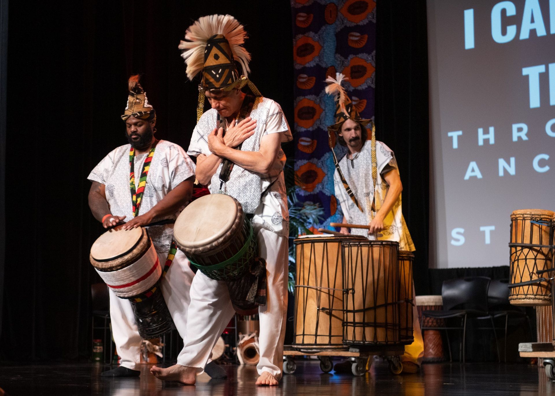Three people in white outfits and headdresses play drums on stage. A banner reads 