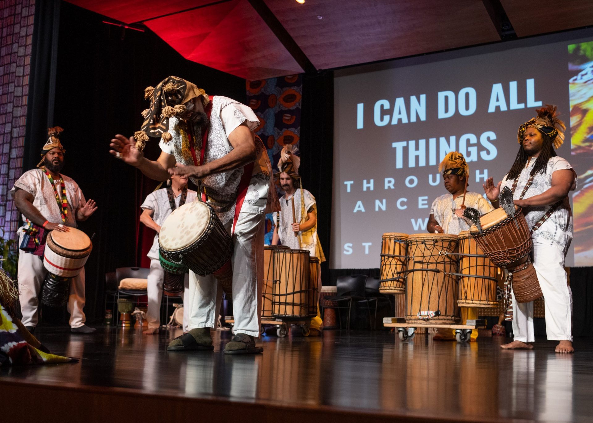 A group of people performing on a stage with drums. Sign reads, 