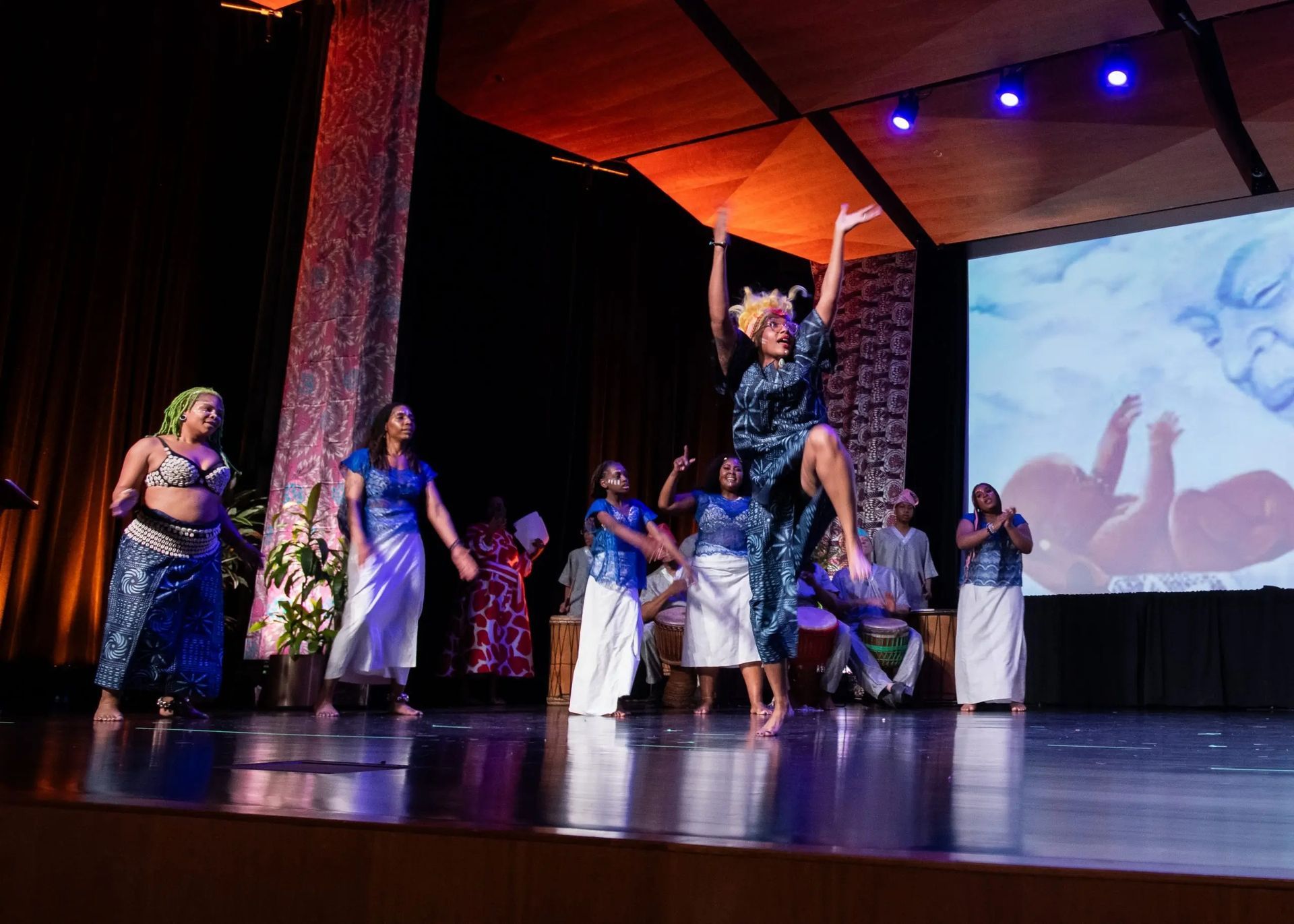 Dancers on stage in blue and white outfits, one leaping with arms raised. Stage with a screen and drums.
