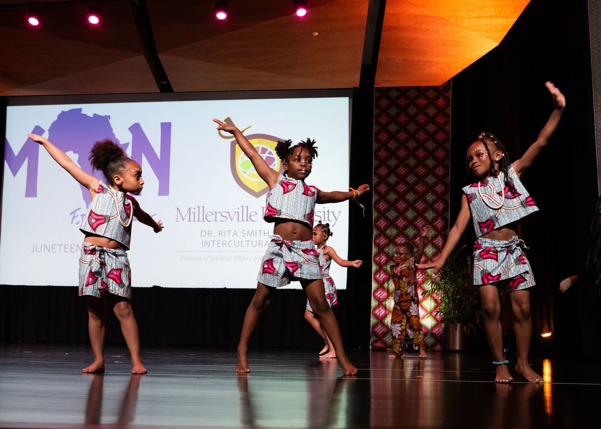 Children dancing on a stage in matching patterned outfits. They have their arms raised, performing.
