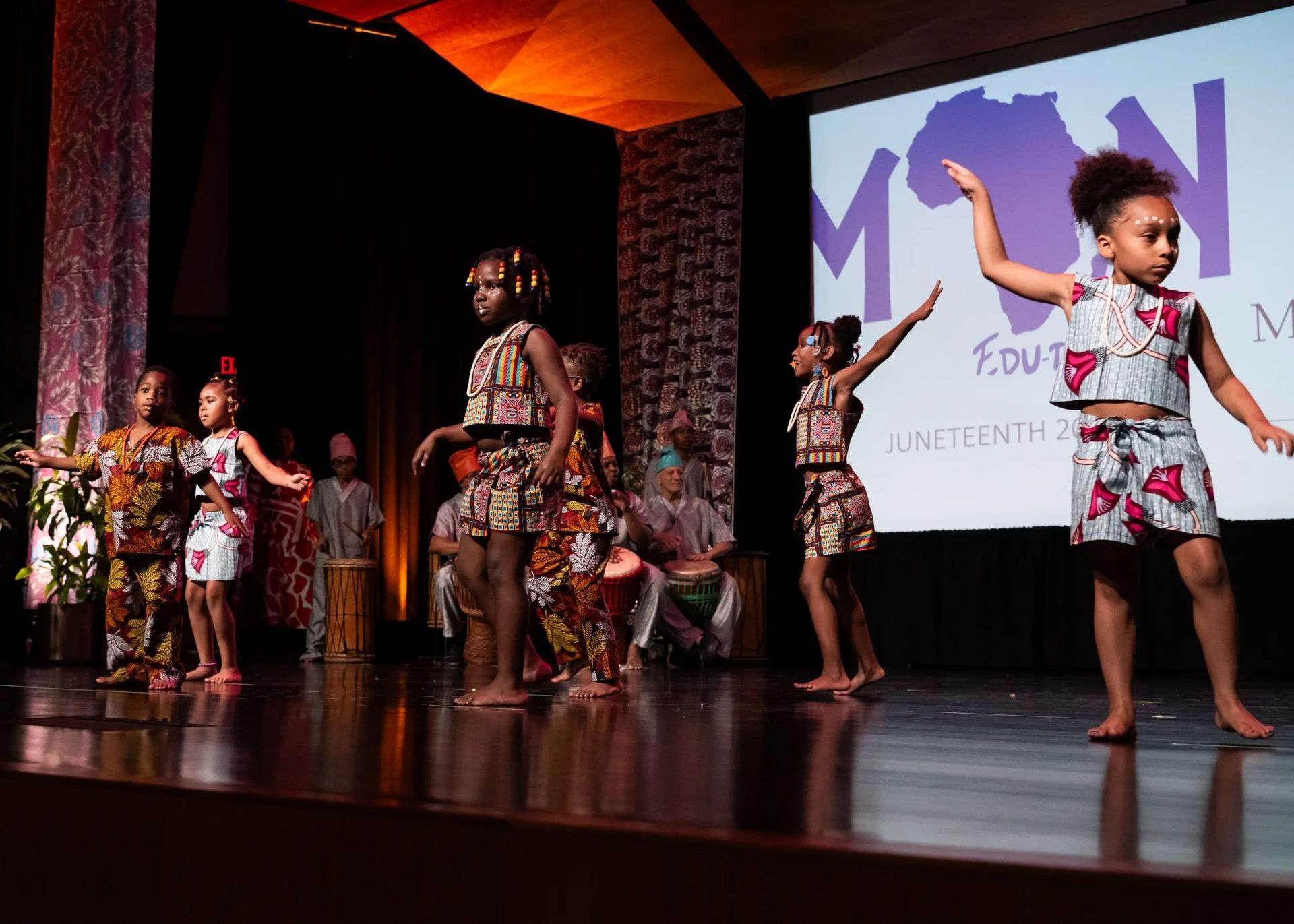 Children in African-print outfits dance on a stage, with a projected logo behind them.