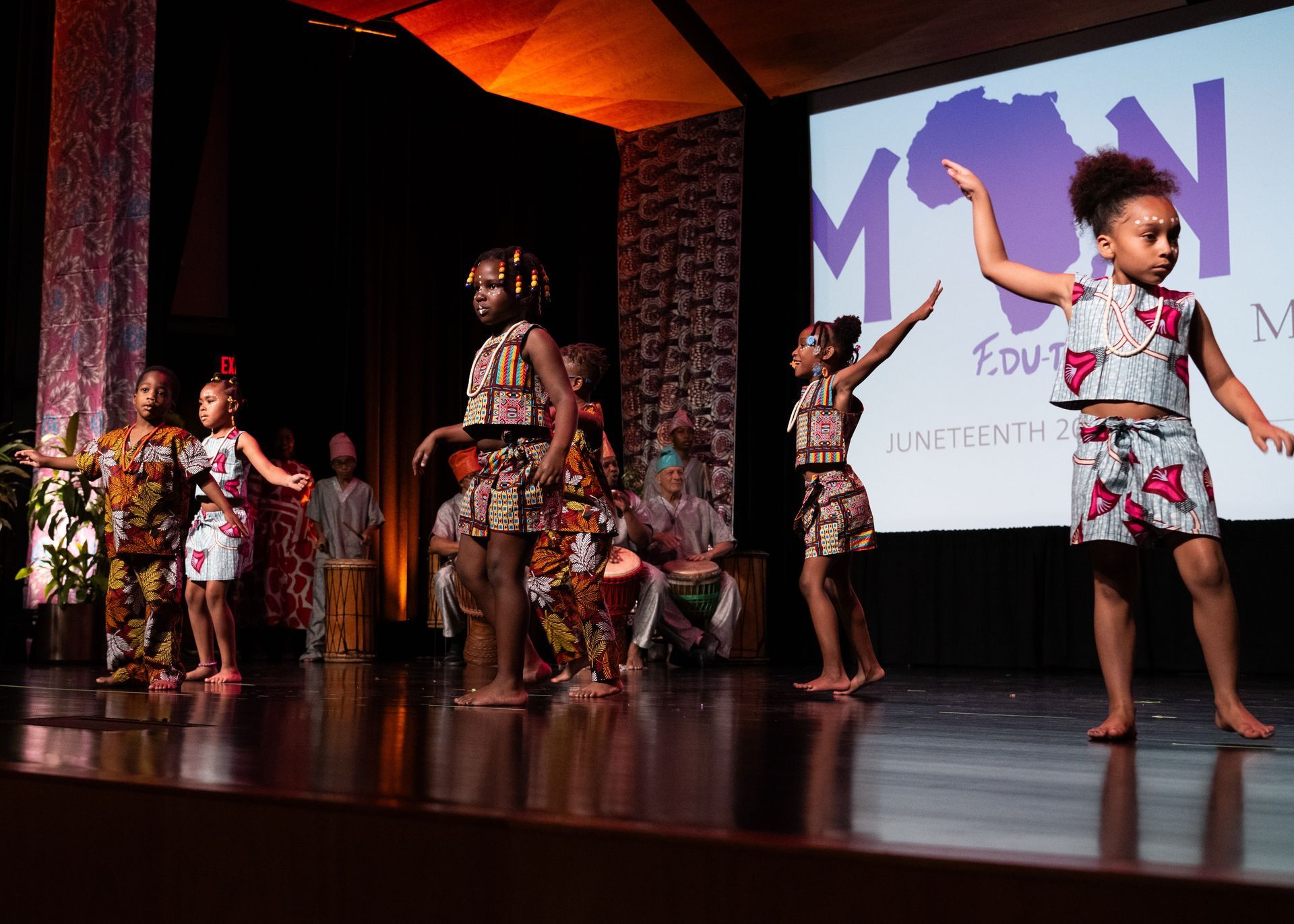 Children in African-print outfits dance on a stage, with a screen displaying a logo and drummers in the background.