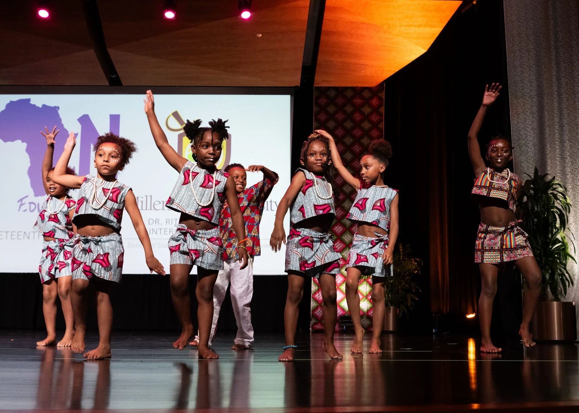 Children in African-print outfits dance on a stage, arms raised. Event backdrop with logo.