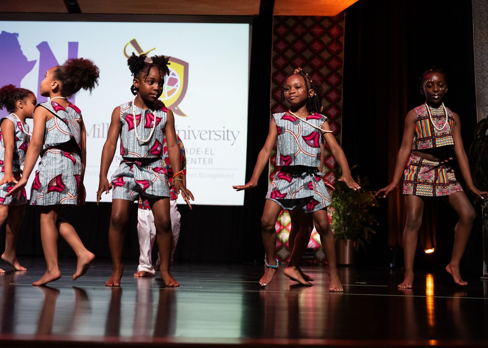 Young girls in African print outfits dancing on a stage with a projected logo.
