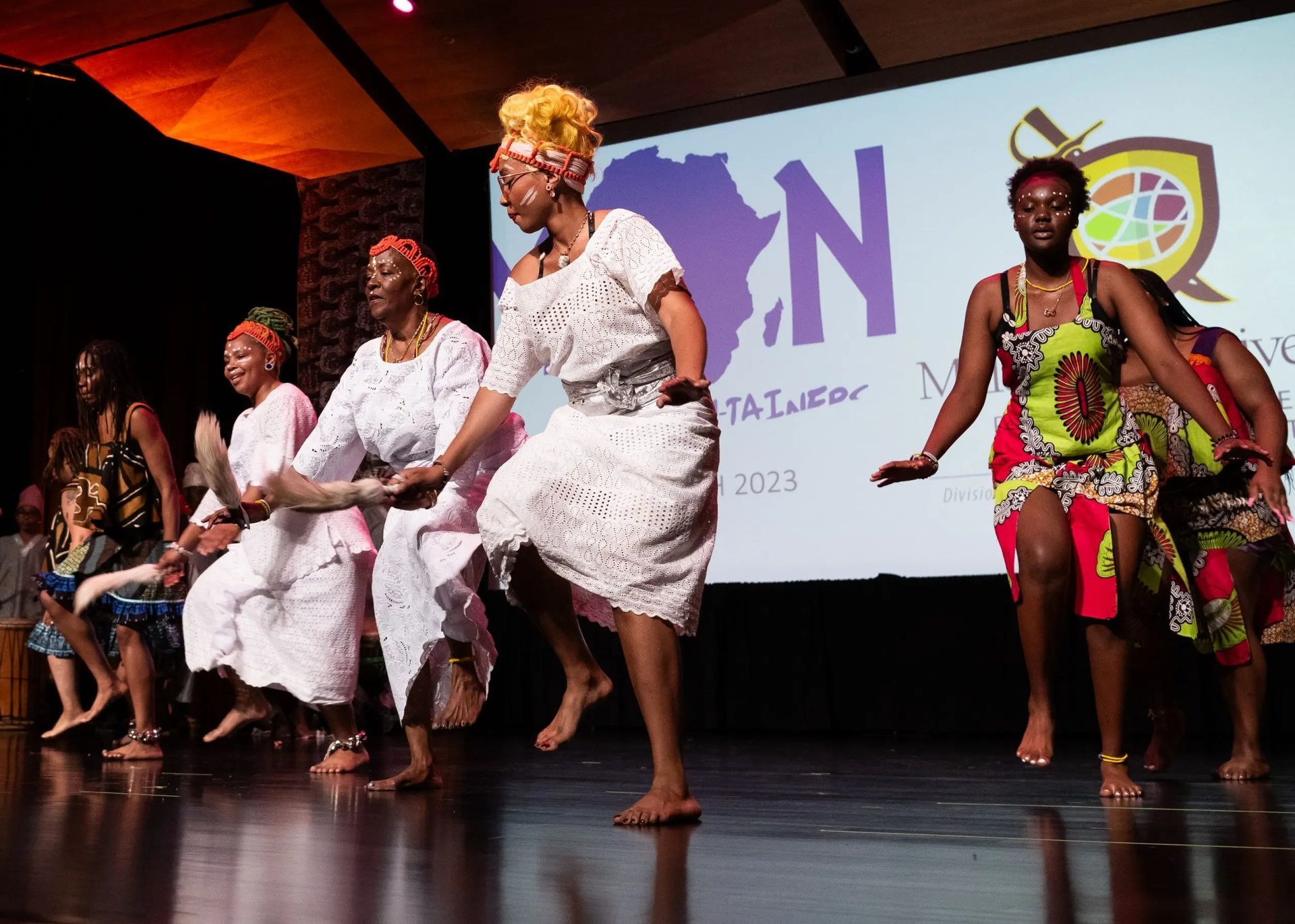 Group of Black women dancing on a stage, wearing colorful and white traditional attire. Stage has a projected backdrop.