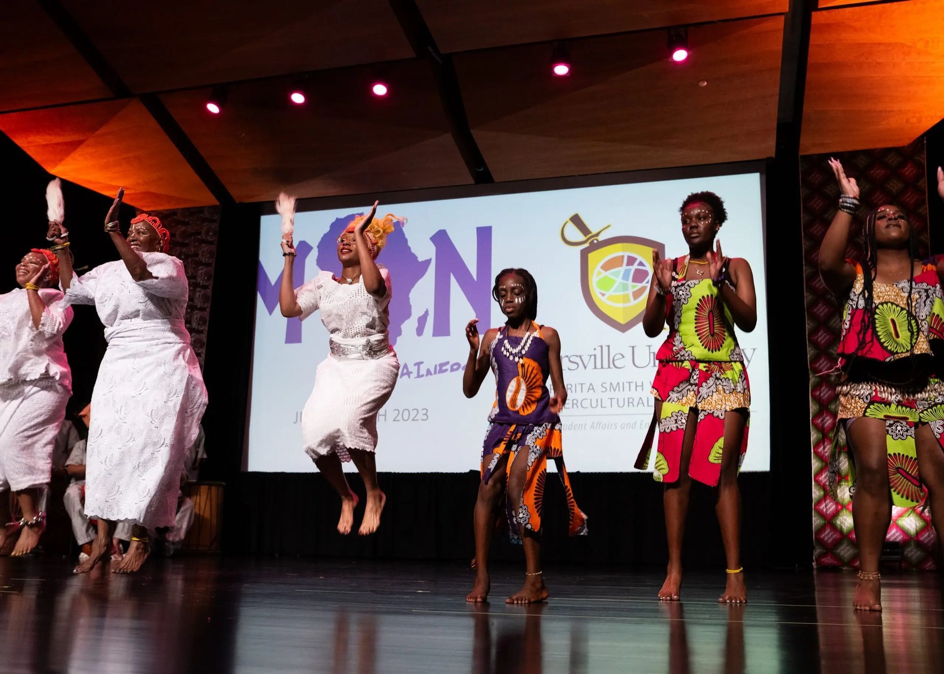 African dancers in colorful attire perform on stage with raised arms.