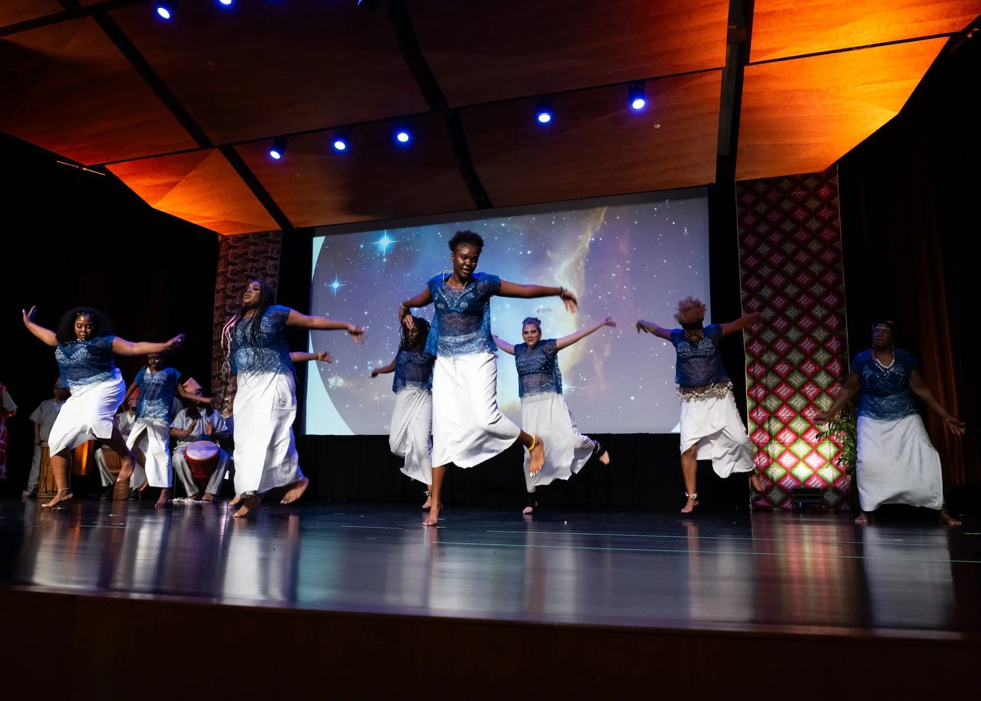 Group of dancers in blue tops and white skirts on a stage, performing in front of a screen with galaxy background.