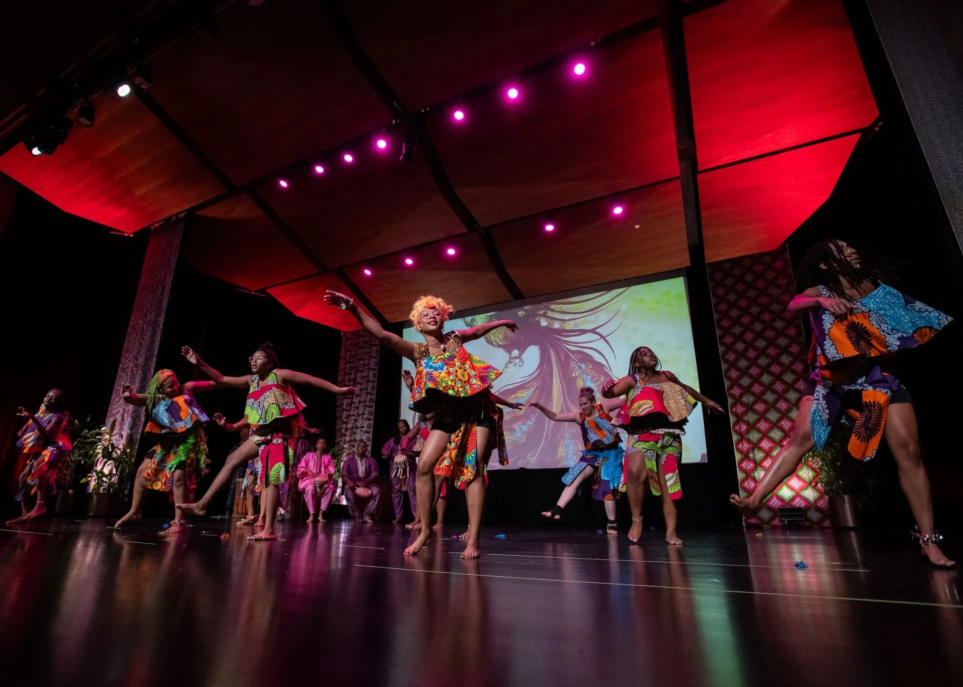 Dancers in colorful African-print costumes perform on stage. Backdrop shows a woman's image.