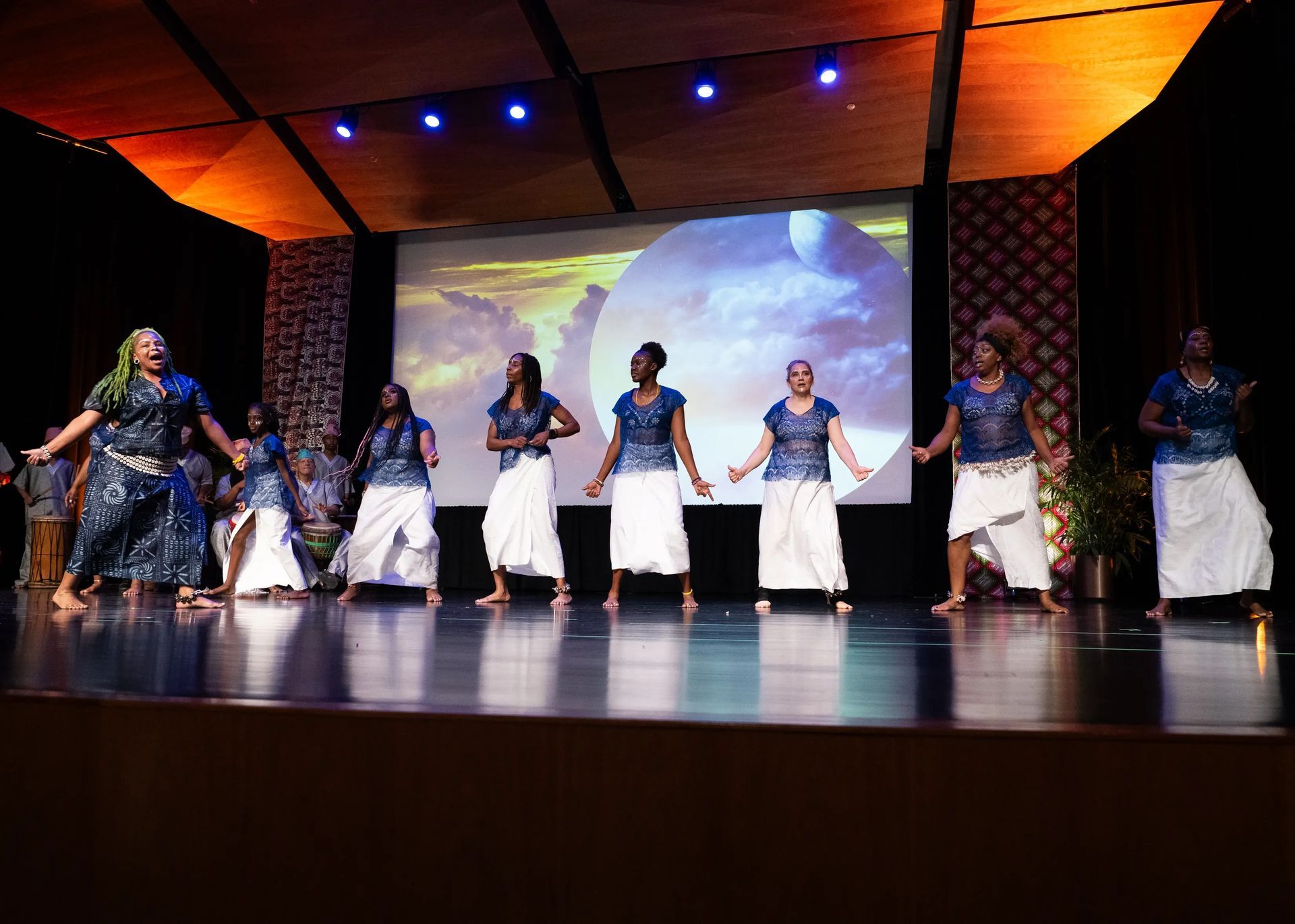 Group of women in blue tops and white skirts dancing on stage.