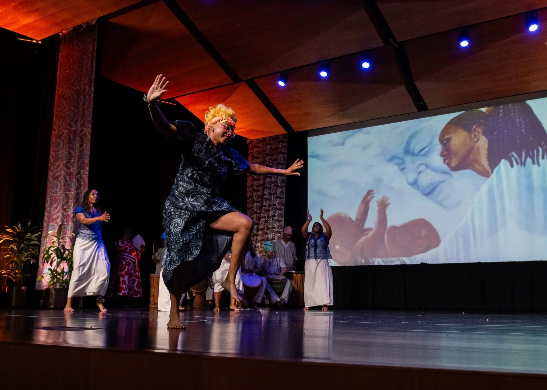 Dancer on stage with others, arms raised, in front of a projected painting of a Black mother and baby.