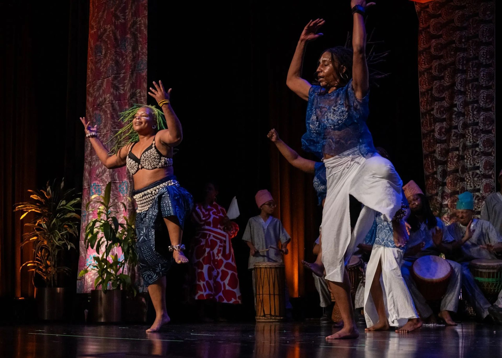 Two dancers in blue costumes perform on stage, with musicians and audience visible.