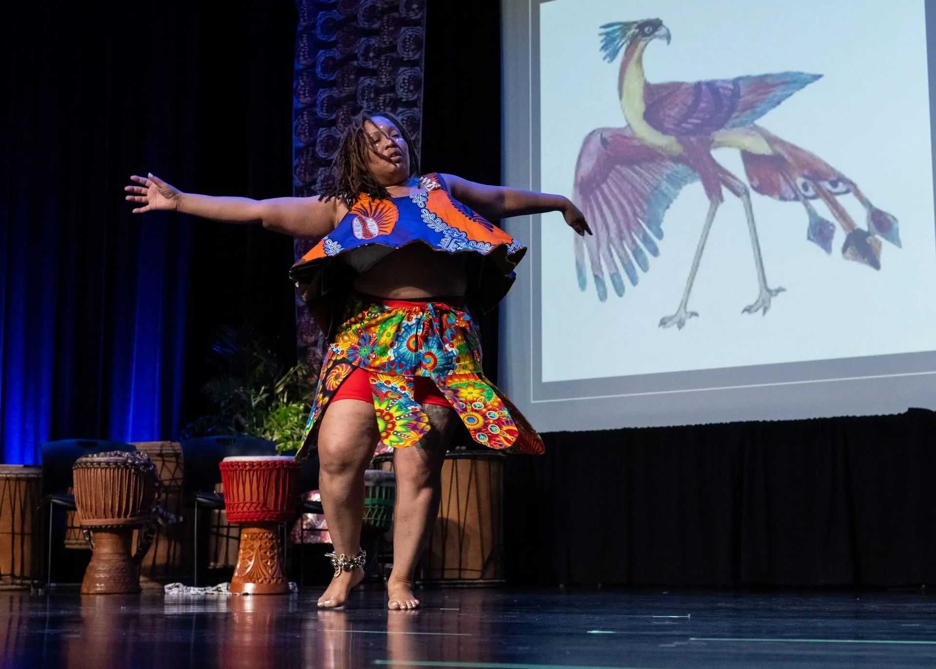 Woman dancing on stage in colorful African attire; projection of a bird behind her.