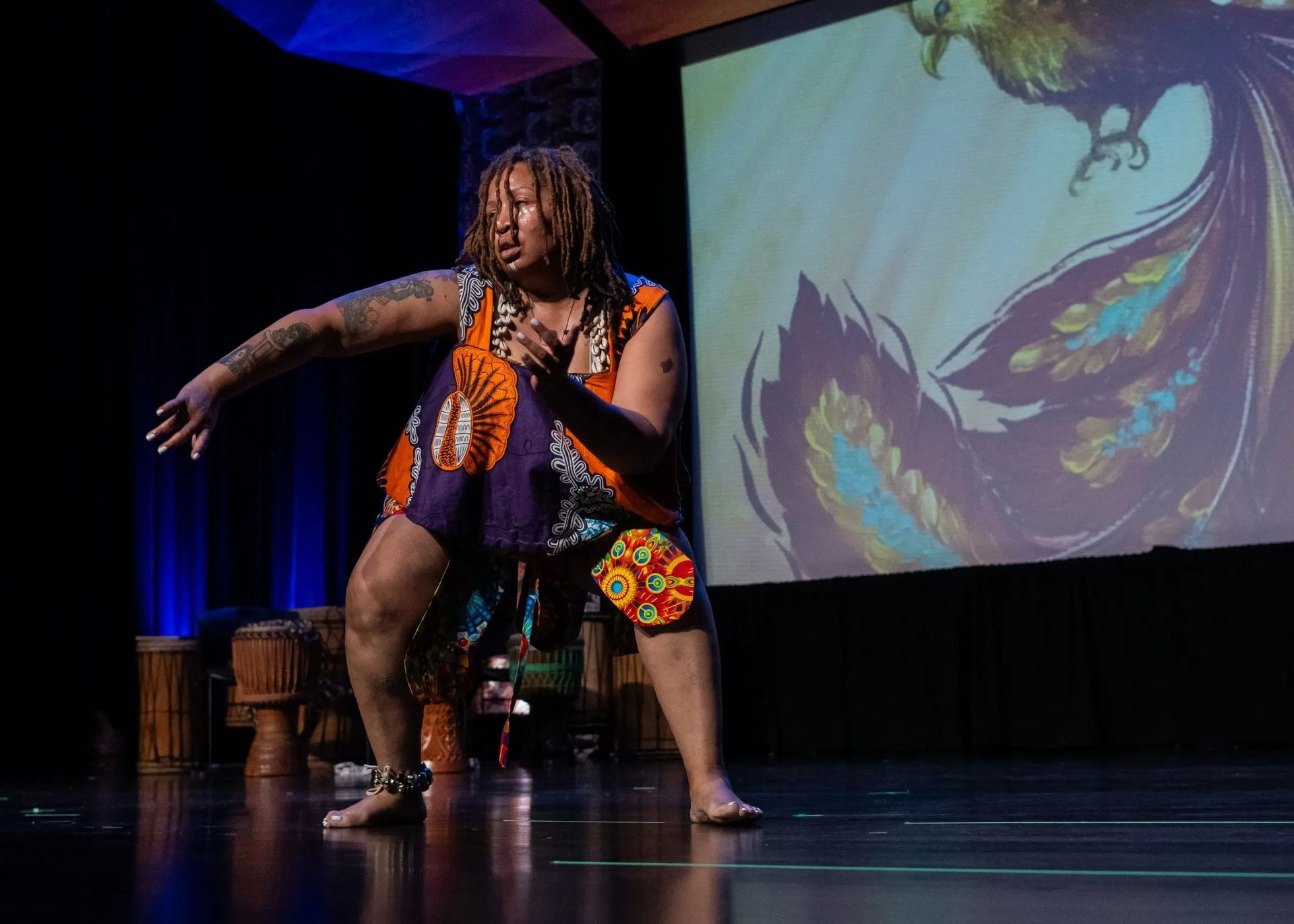 Person in vibrant African attire dances on stage with a mural background.