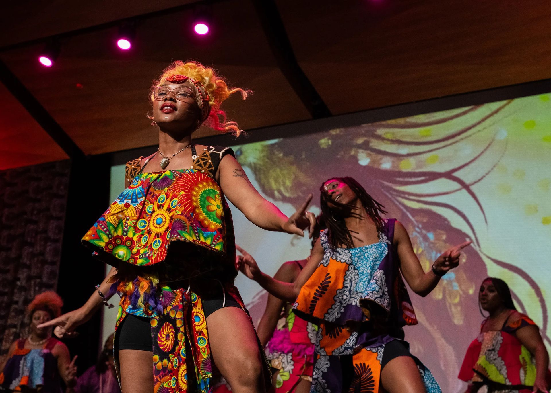Dancers in colorful African-print outfits on stage with a projected backdrop, performing with energy.