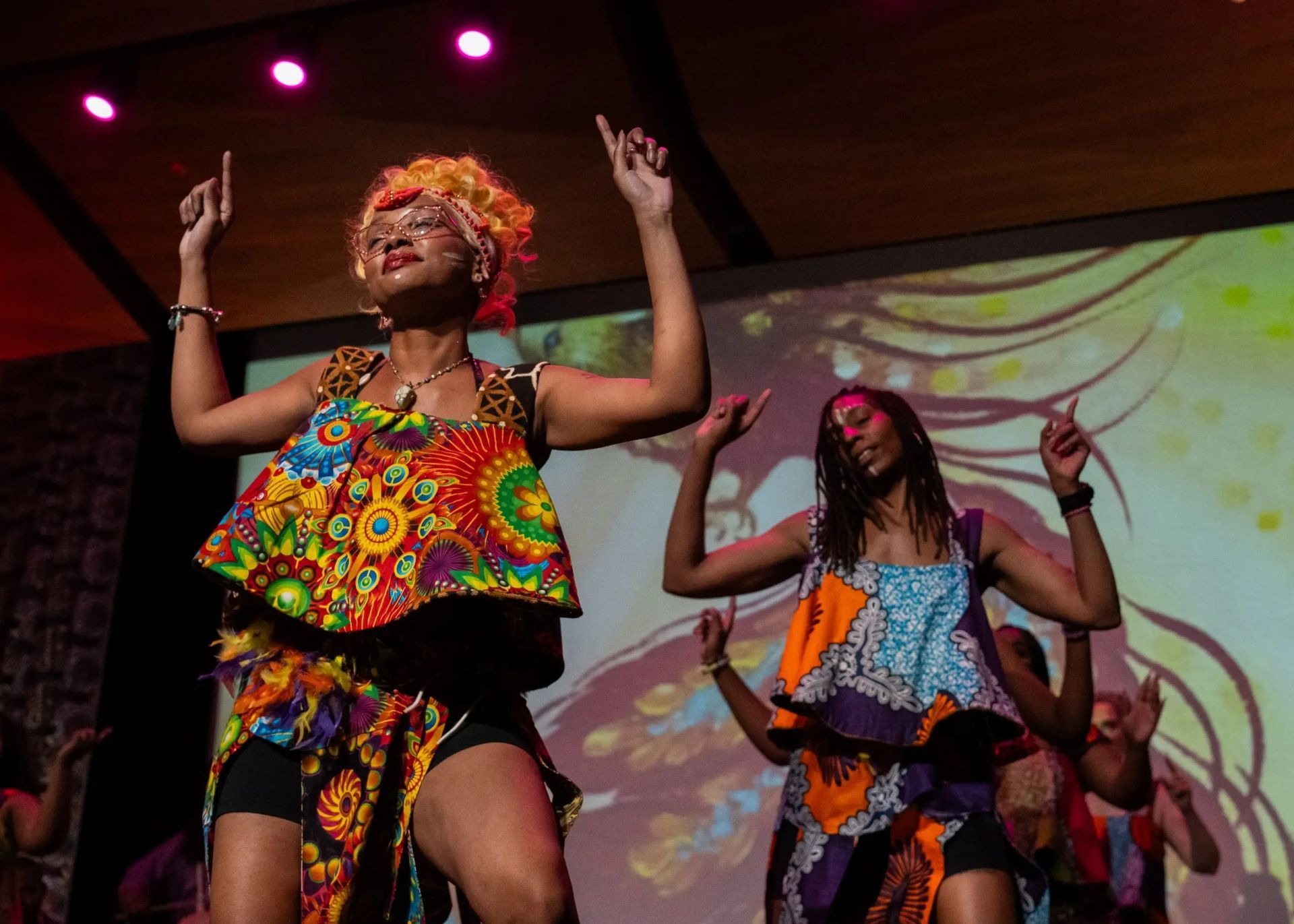 Two Black women dancing on stage in vibrant African print outfits, arms raised.