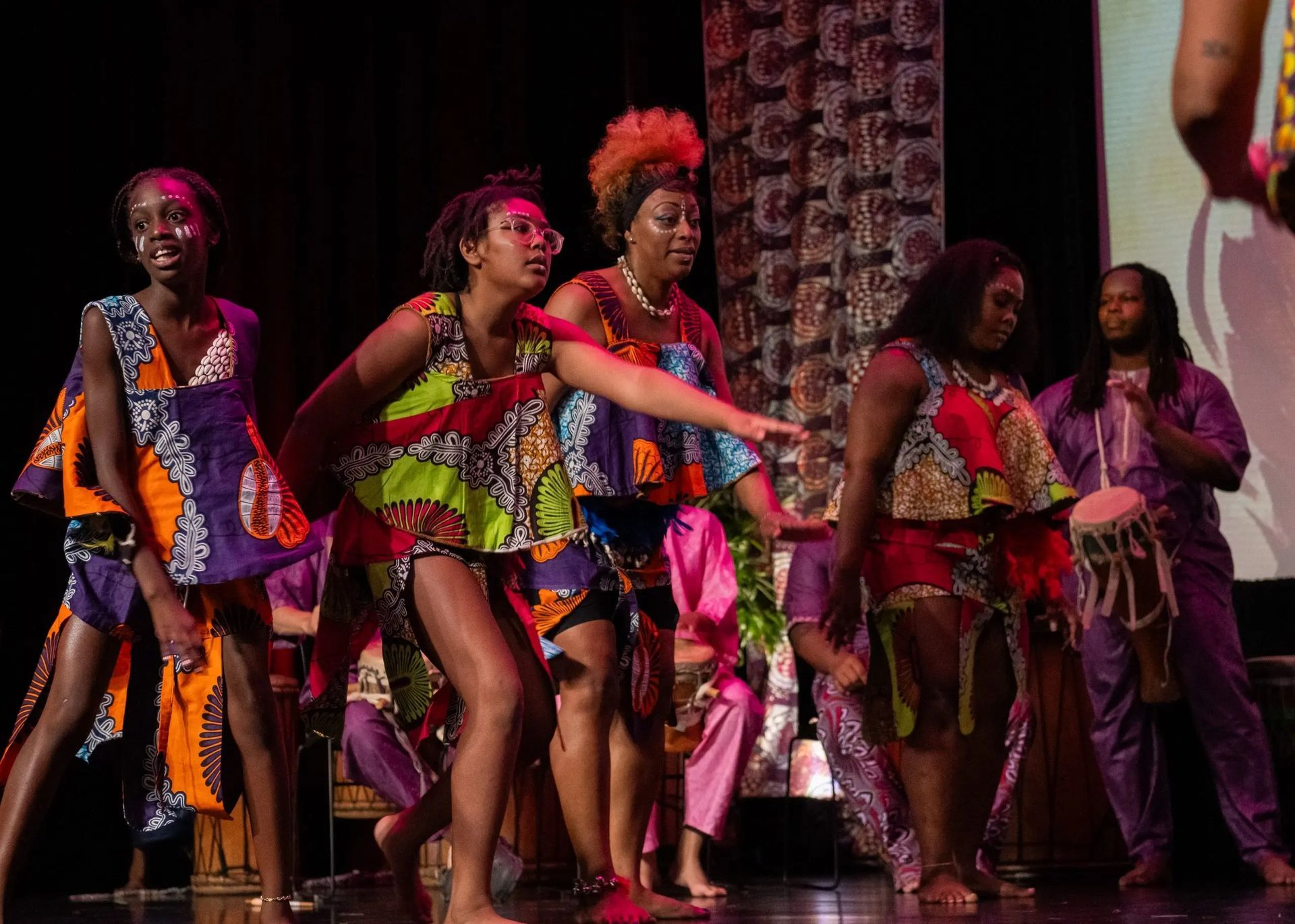 Dancers in colorful African print costumes on stage. Woman in the center gestures. Drummer at right.