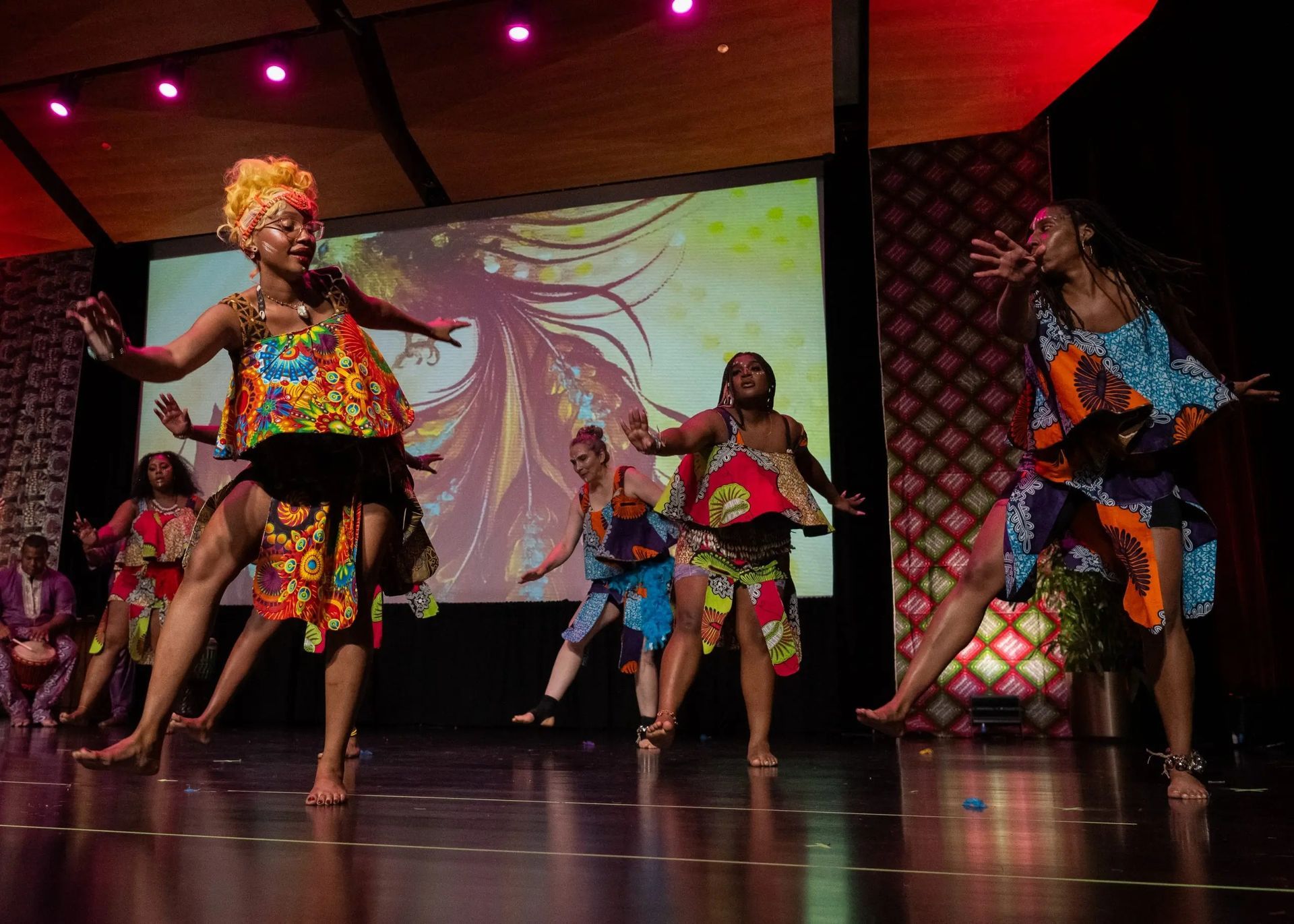African dancers in colorful patterned clothing perform on stage under bright lights.