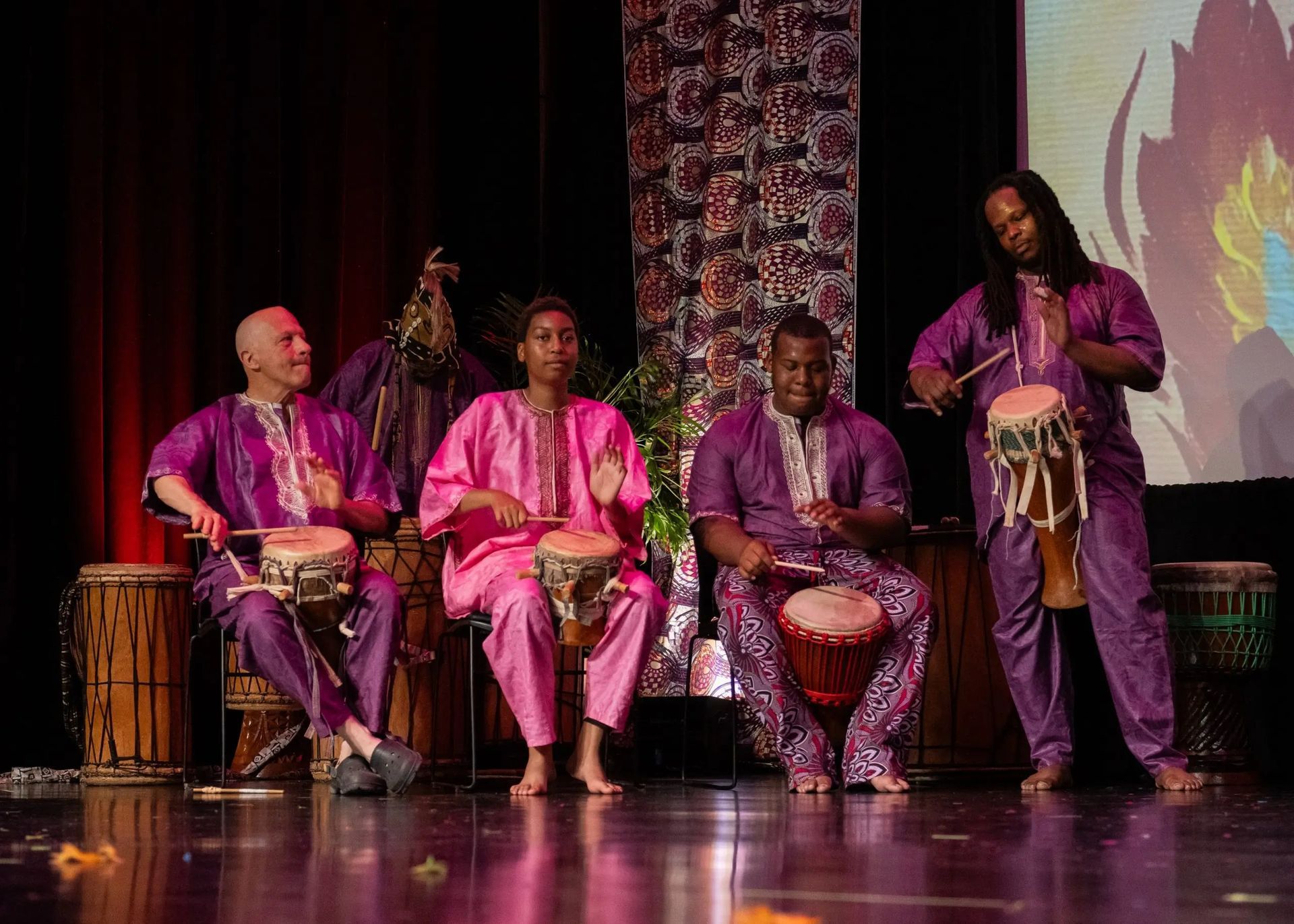 Five people in purple and pink outfits play drums on a stage with a decorative backdrop.