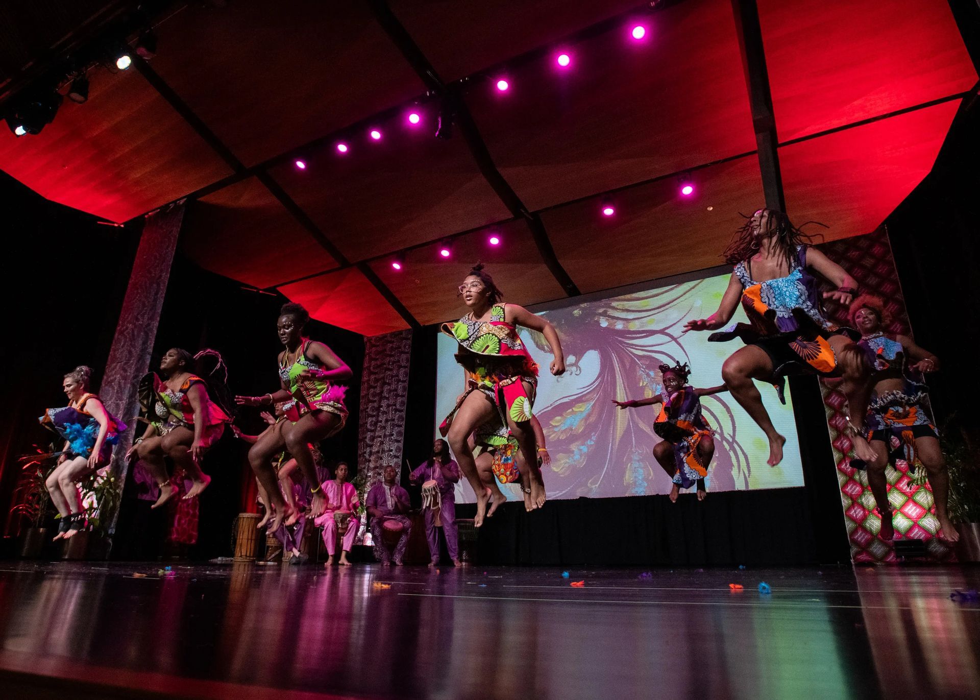 Dancers in colorful costumes jump on stage under red lights. A screen with a graphic is behind them.