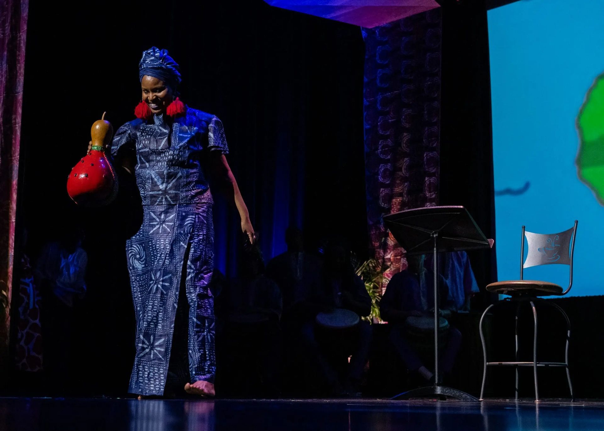 Woman in blue outfit, holding a red gourd, smiles on stage. Dark room, chair, screen.