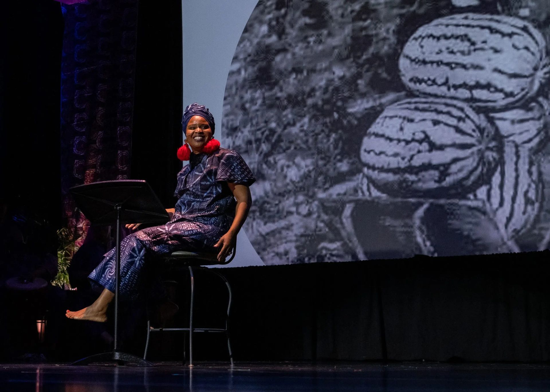 Woman in patterned blue outfit sits on a stool, speaking at a lectern in front of a black-and-white watermelon image.