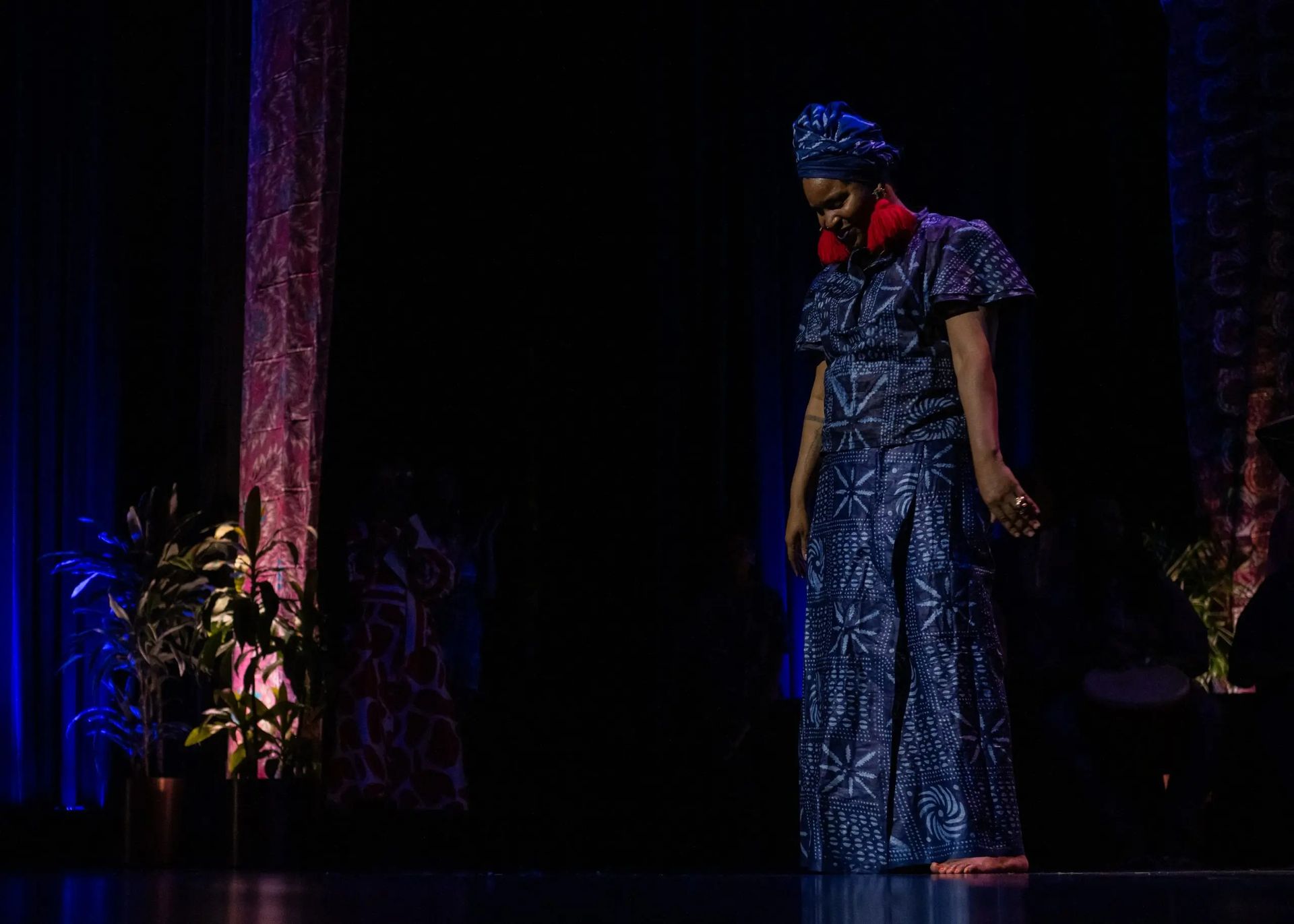 Woman on stage in blue African print dress, head wrap, and red earrings. Dark setting with plants and decorative panels.
