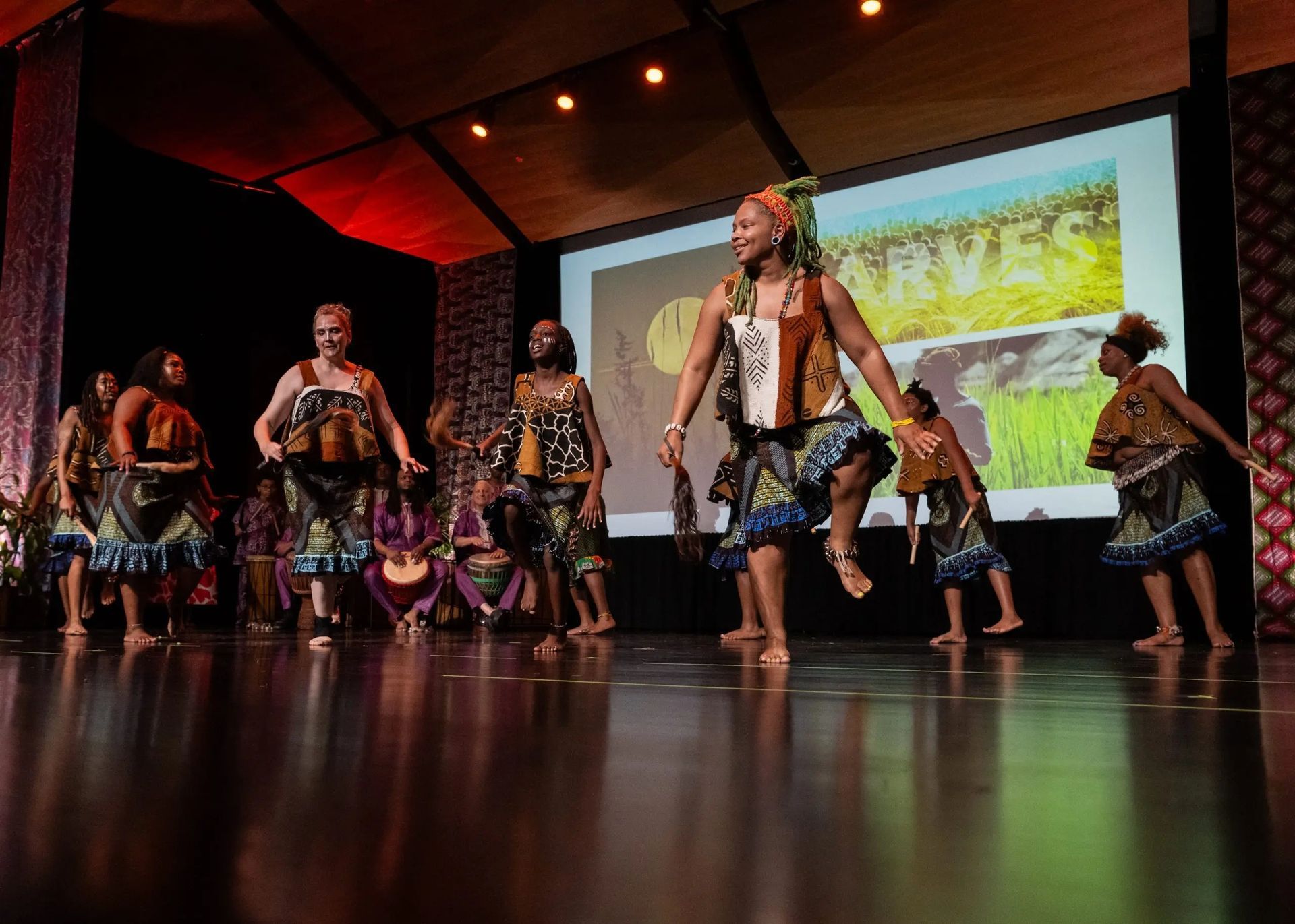 African dancers on a stage. The dancers are performing, wearing colorful costumes, with a backdrop of a landscape.
