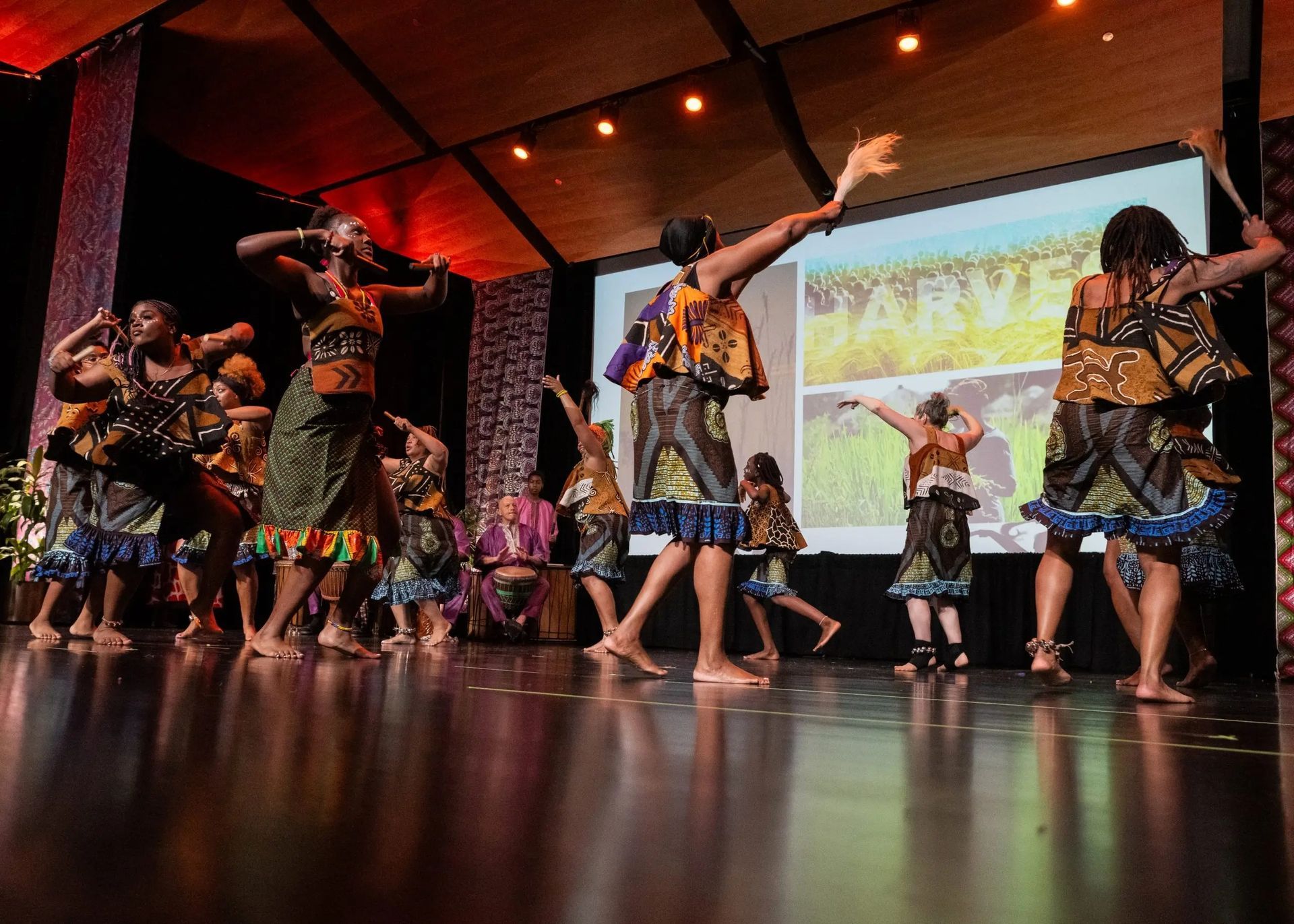 Dancers in colorful costumes perform on stage; a screen in background shows images.