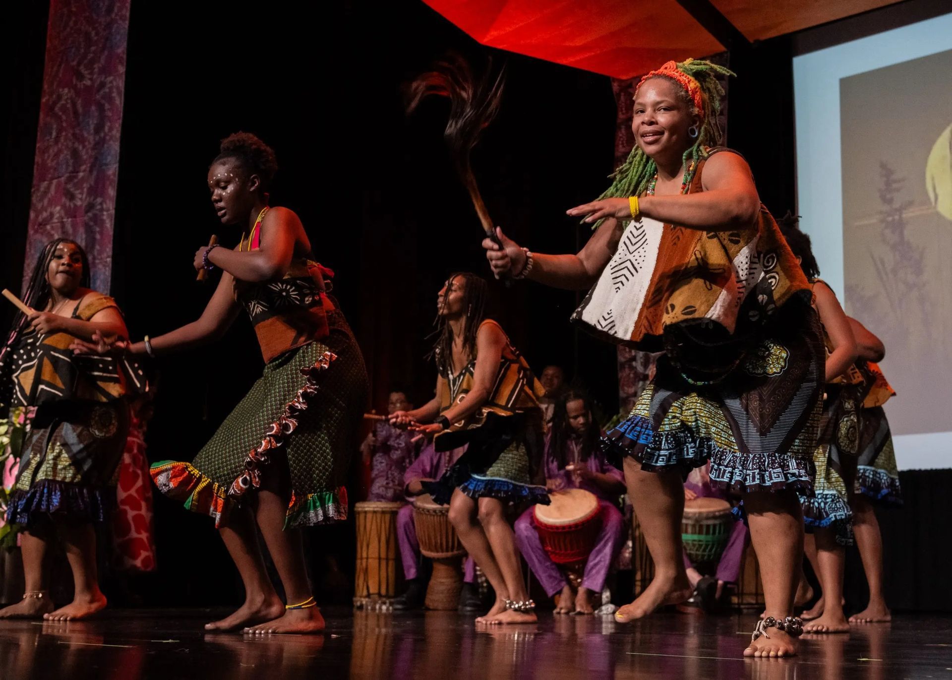 African dancers on stage with drums, wearing colorful patterned clothing, moving joyfully.