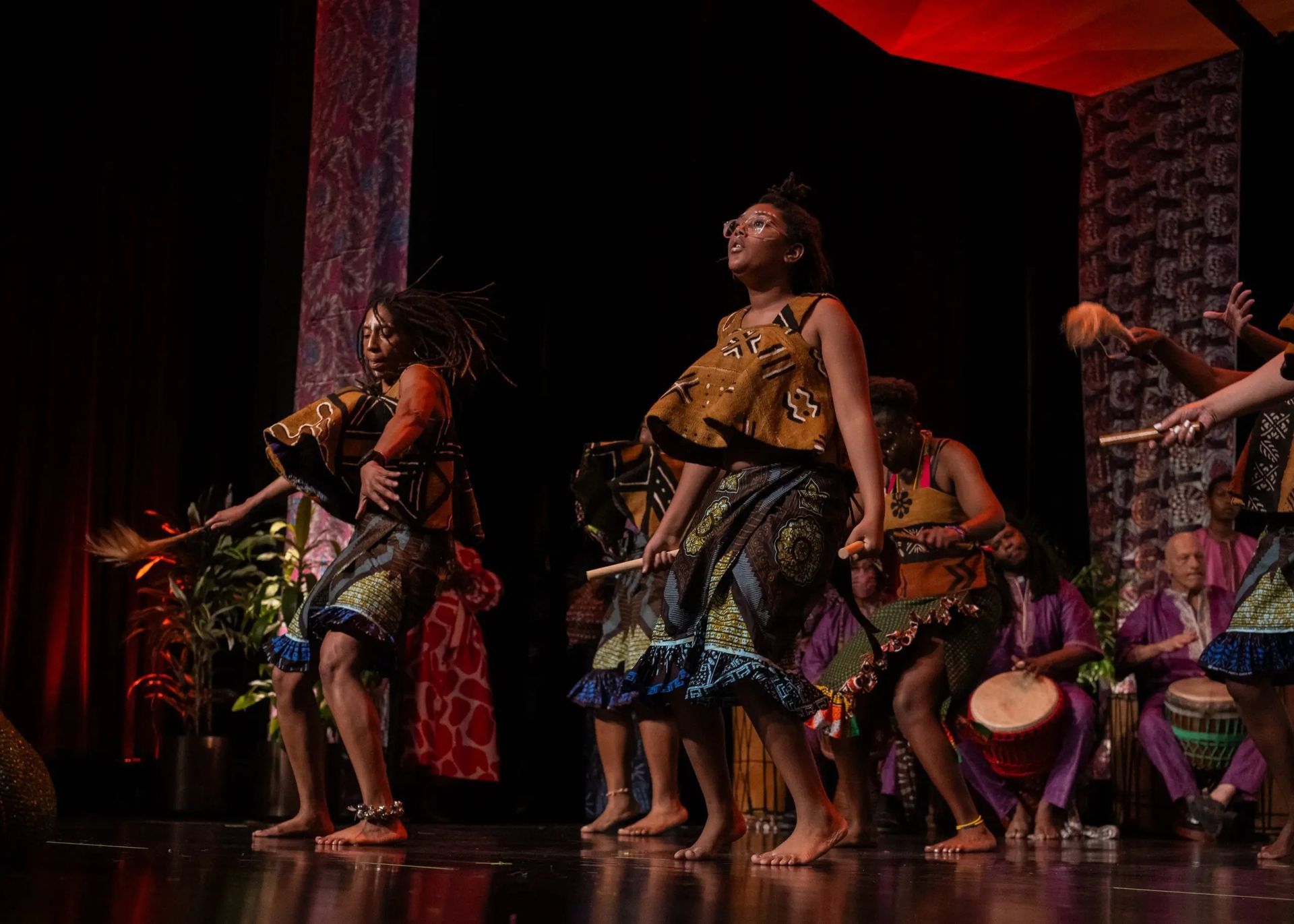 African dancers perform on stage, wearing colorful costumes. Drummers sit in the background.