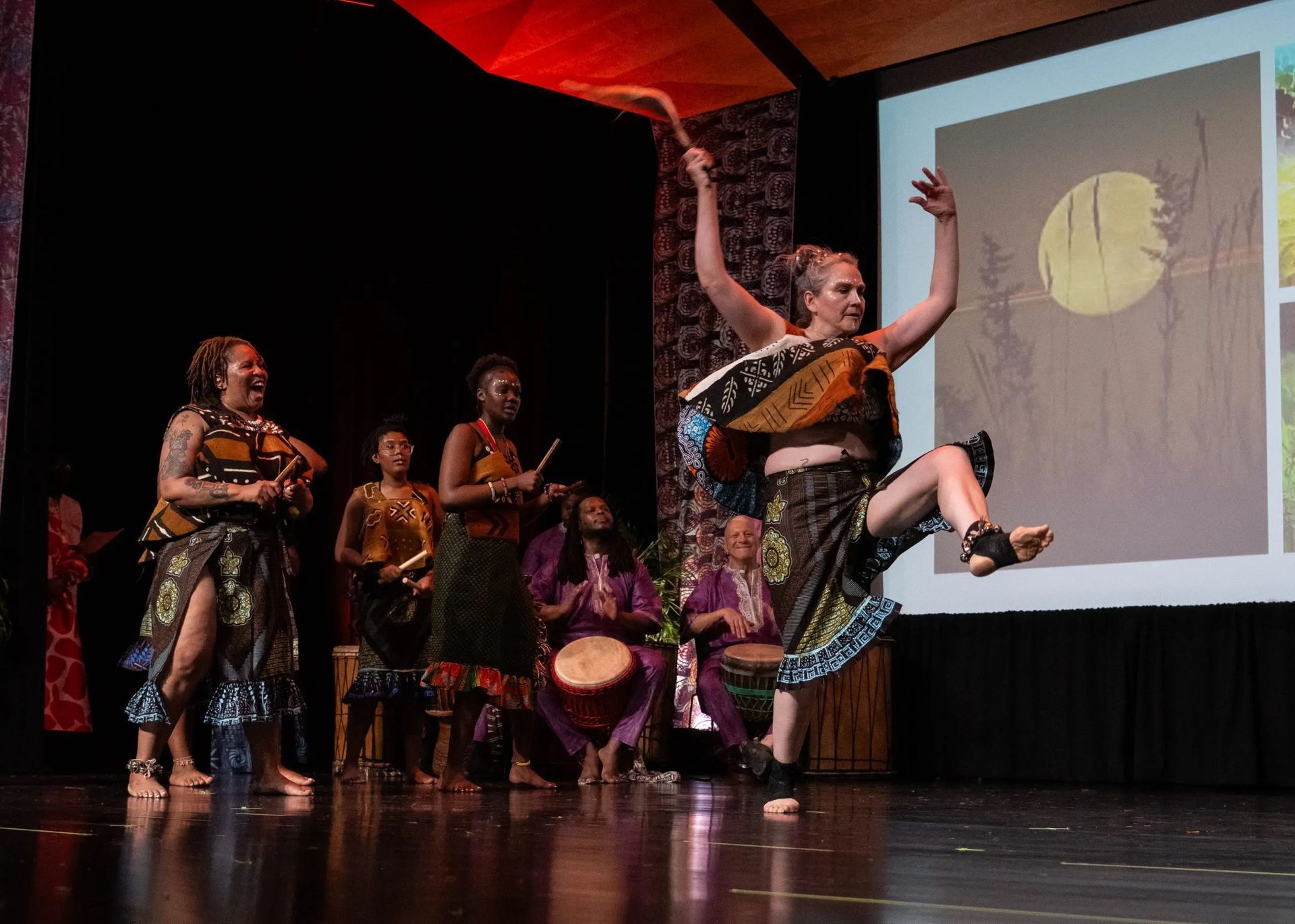 Group of dancers in colorful costumes perform on stage with musical instruments and a projected backdrop.