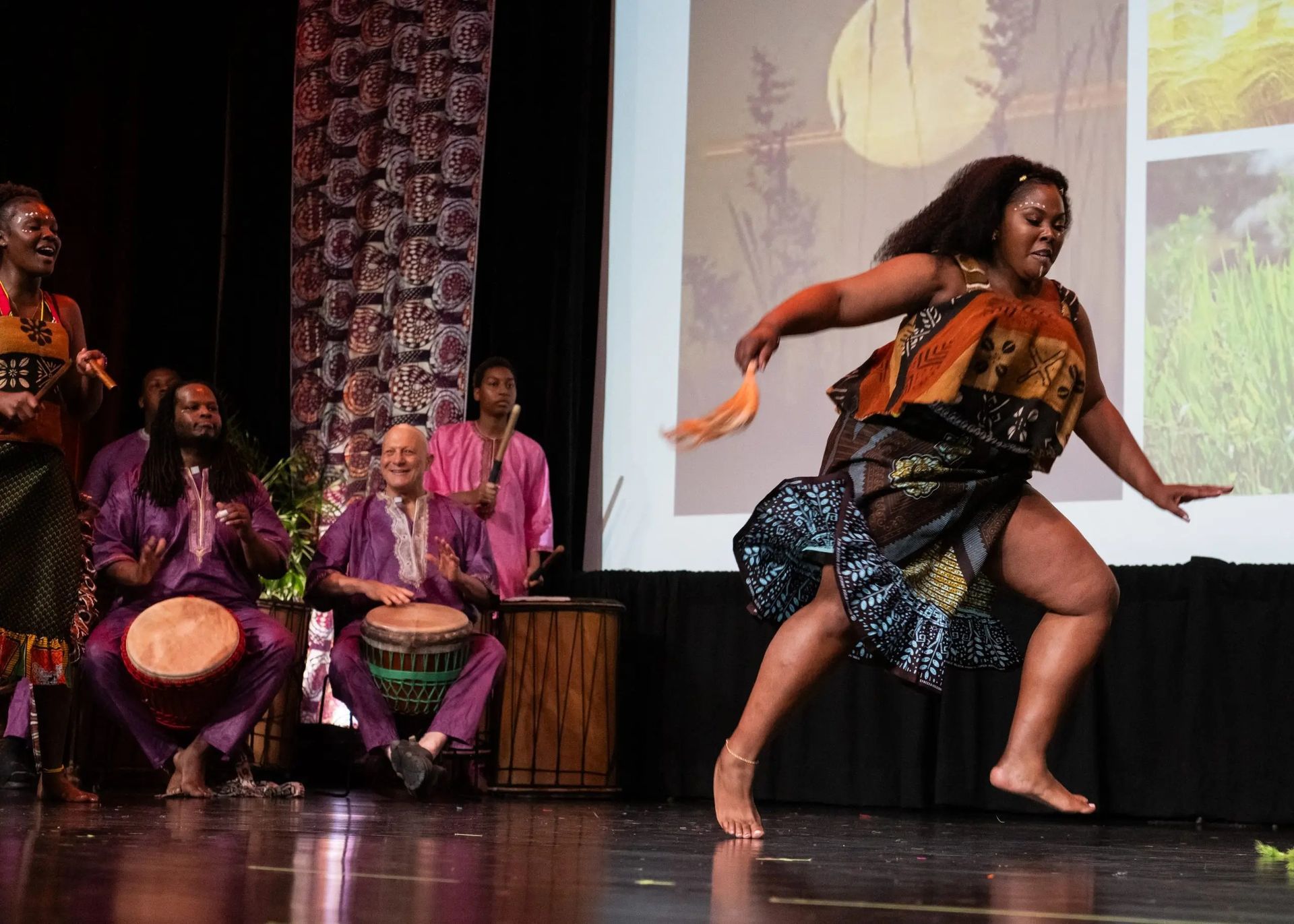 A dancer performs on stage with musicians in colorful outfits; a screen displays nature images.