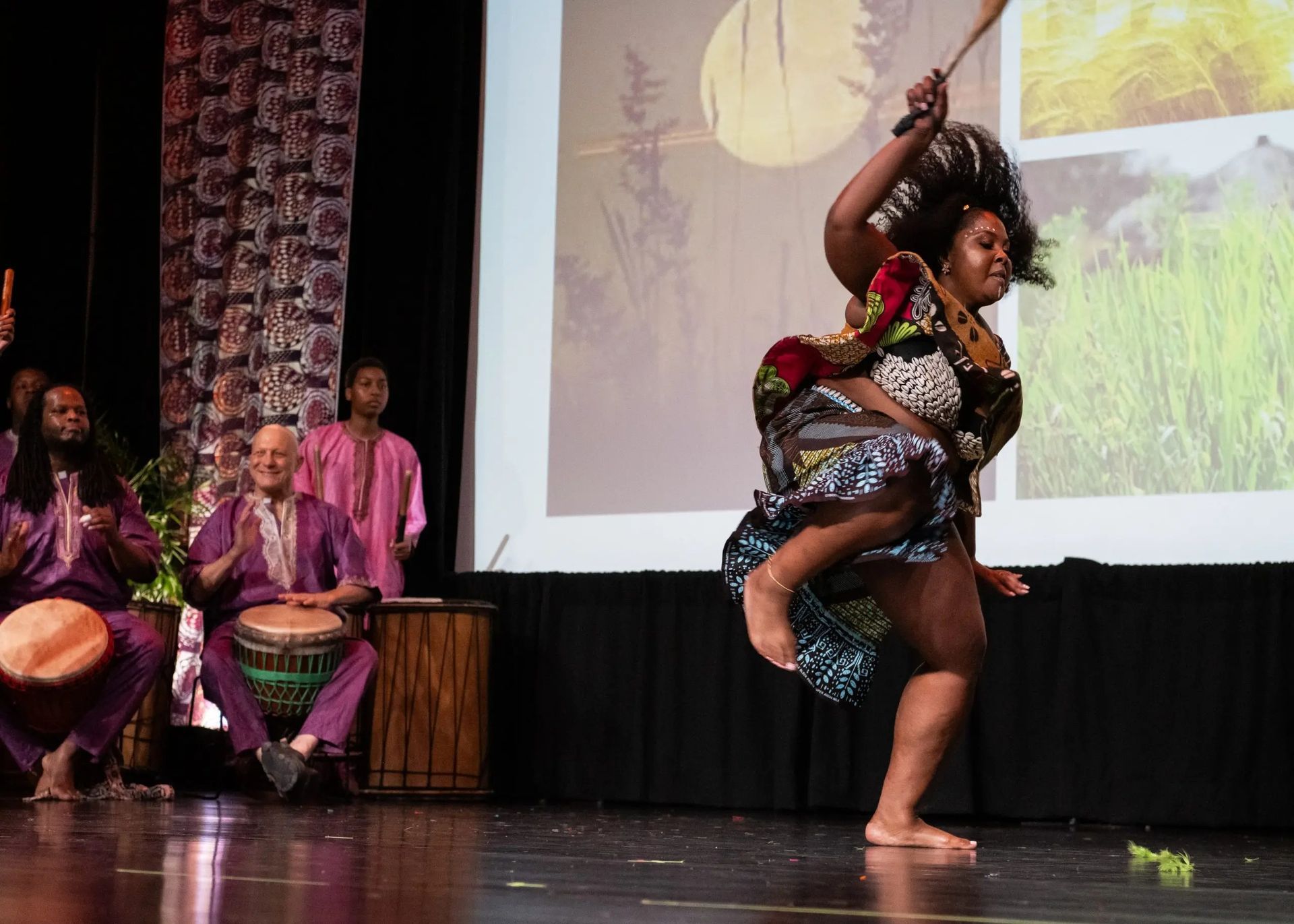 Woman dances on stage with drums, colorful outfit, and raised stick. Men play drums, background projection.