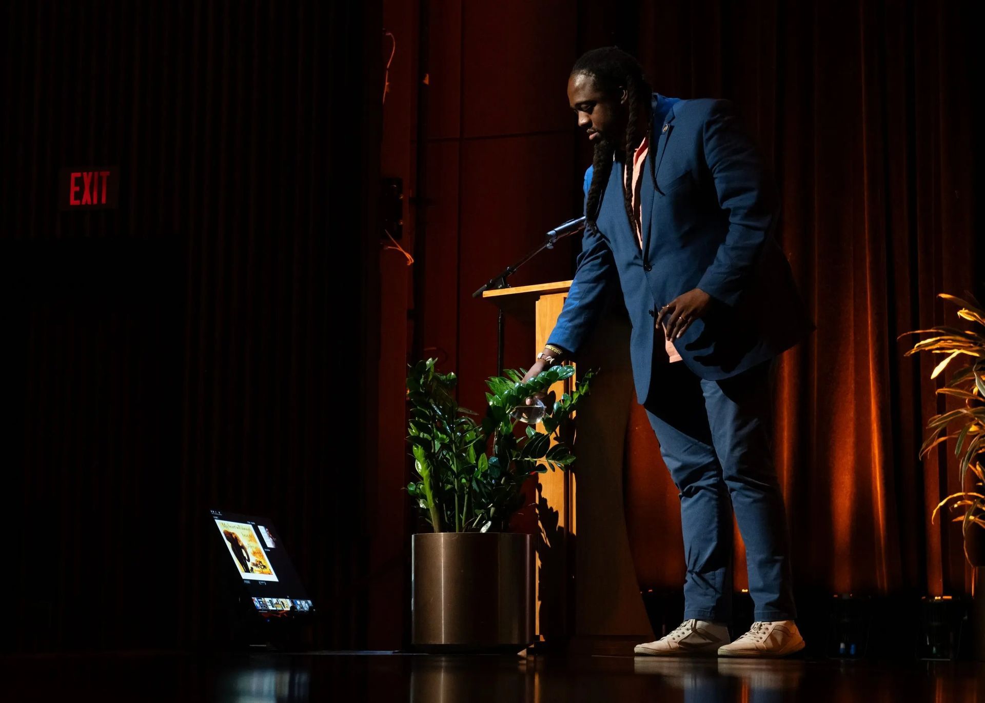 Man in blue suit at a podium reaches toward a plant next to a laptop screen in a dark room.